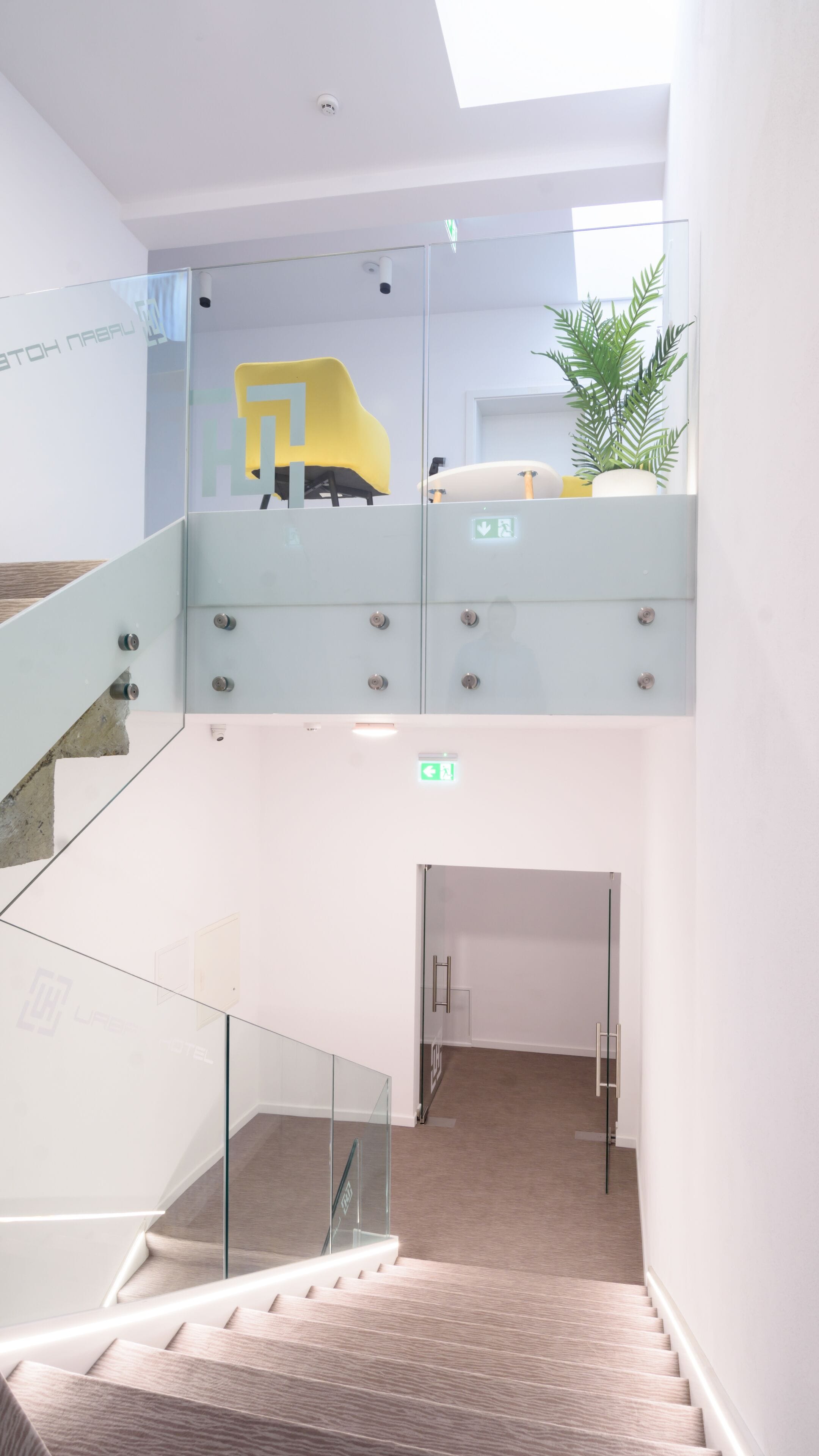 A modern hotel interior with a glass staircase, a yellow chair, and a potted plant.