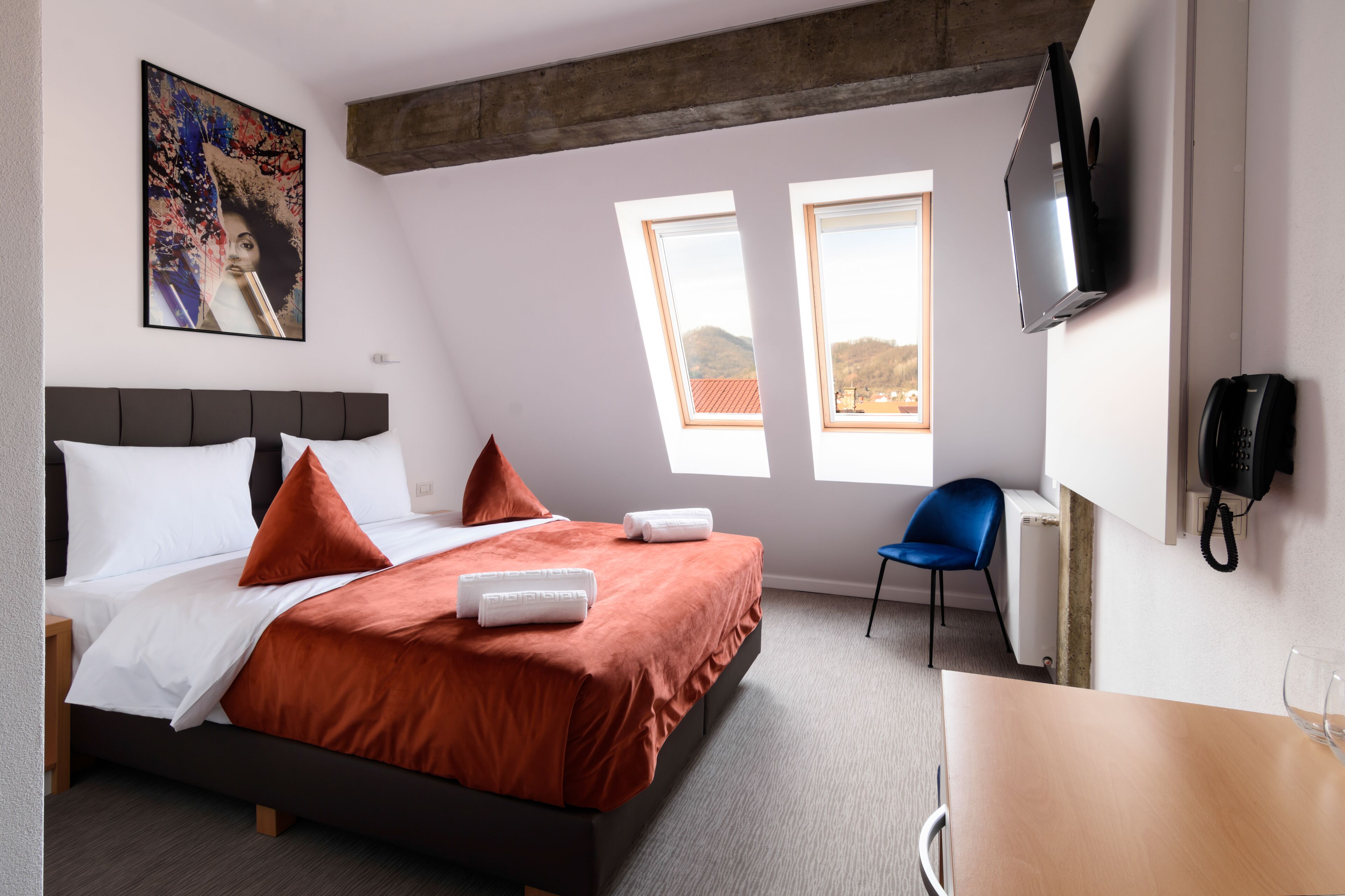 A modern hotel room with a bed, a desk, a chair, a TV, and a painting on the wall.