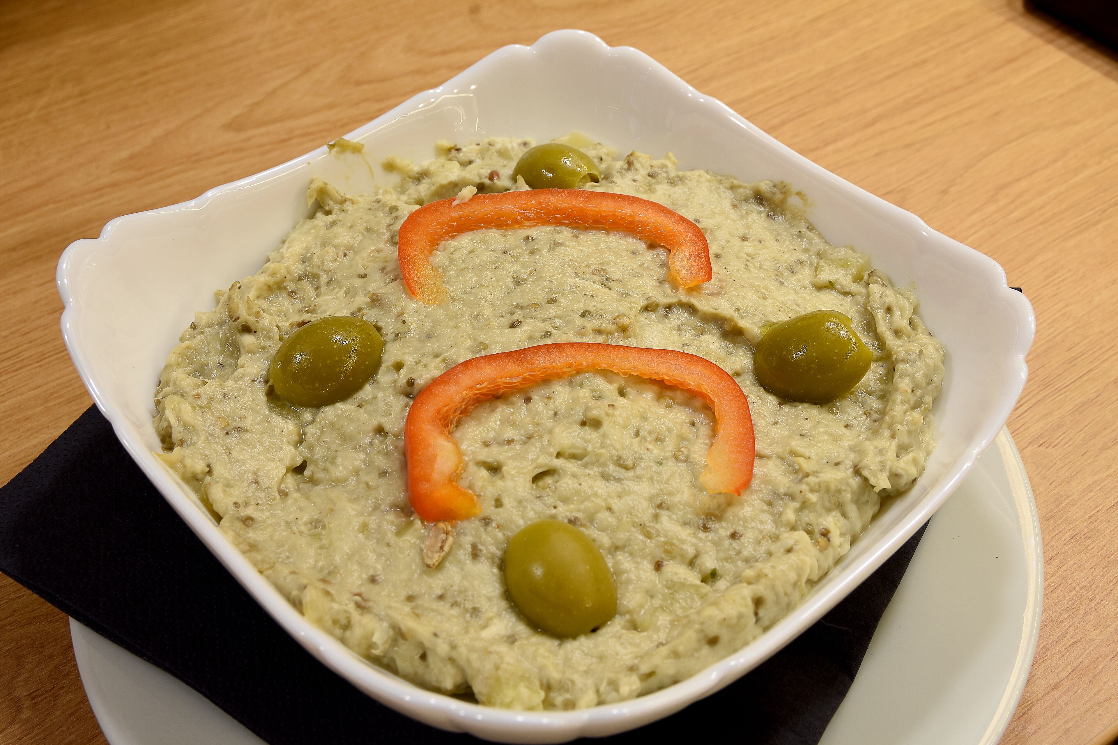 A bowl of dip with olives and a carrot slice on a white plate.