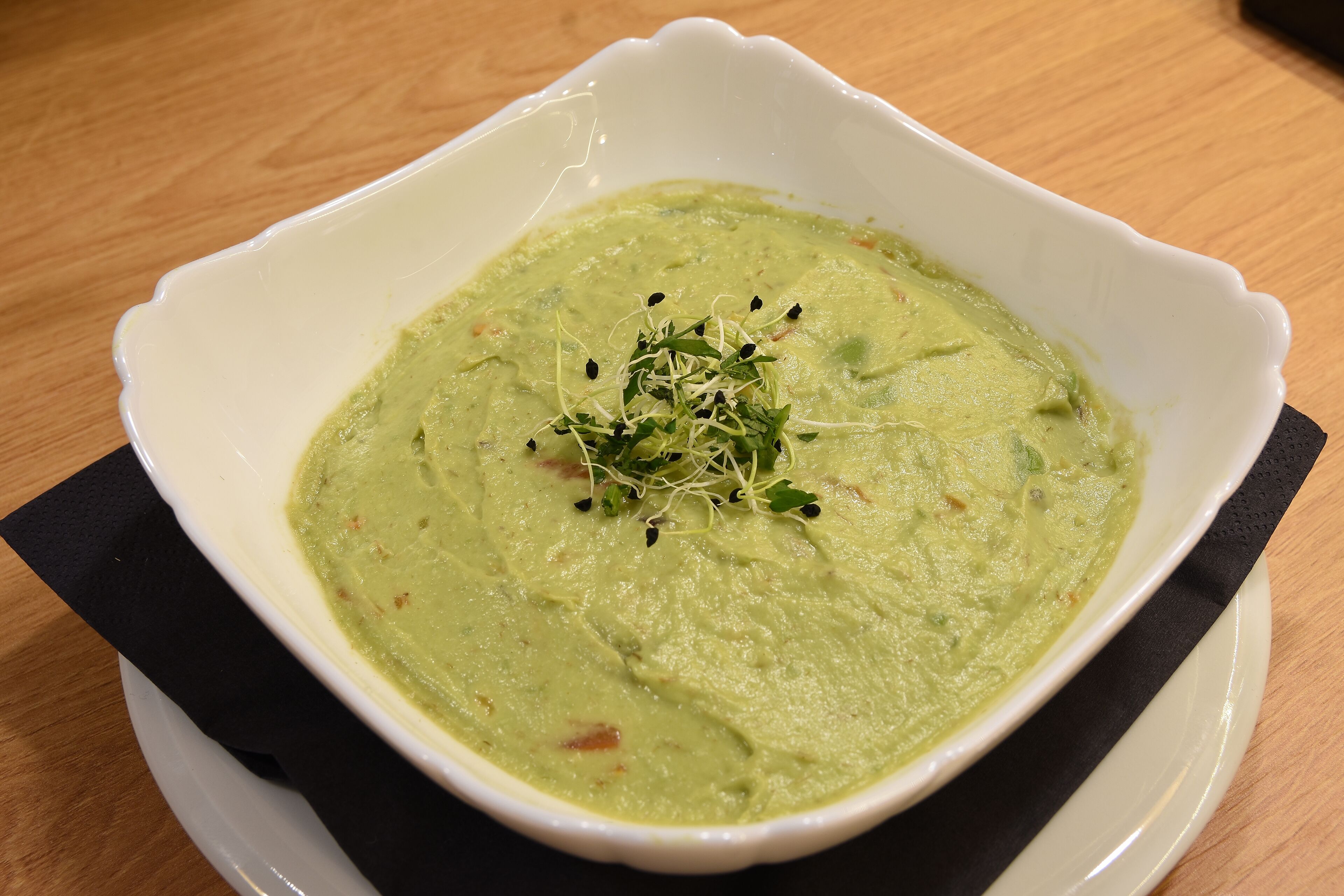 Un tazón blanco con sopa verde y brotes sobre un mantel negro, colocado sobre una mesa de madera.