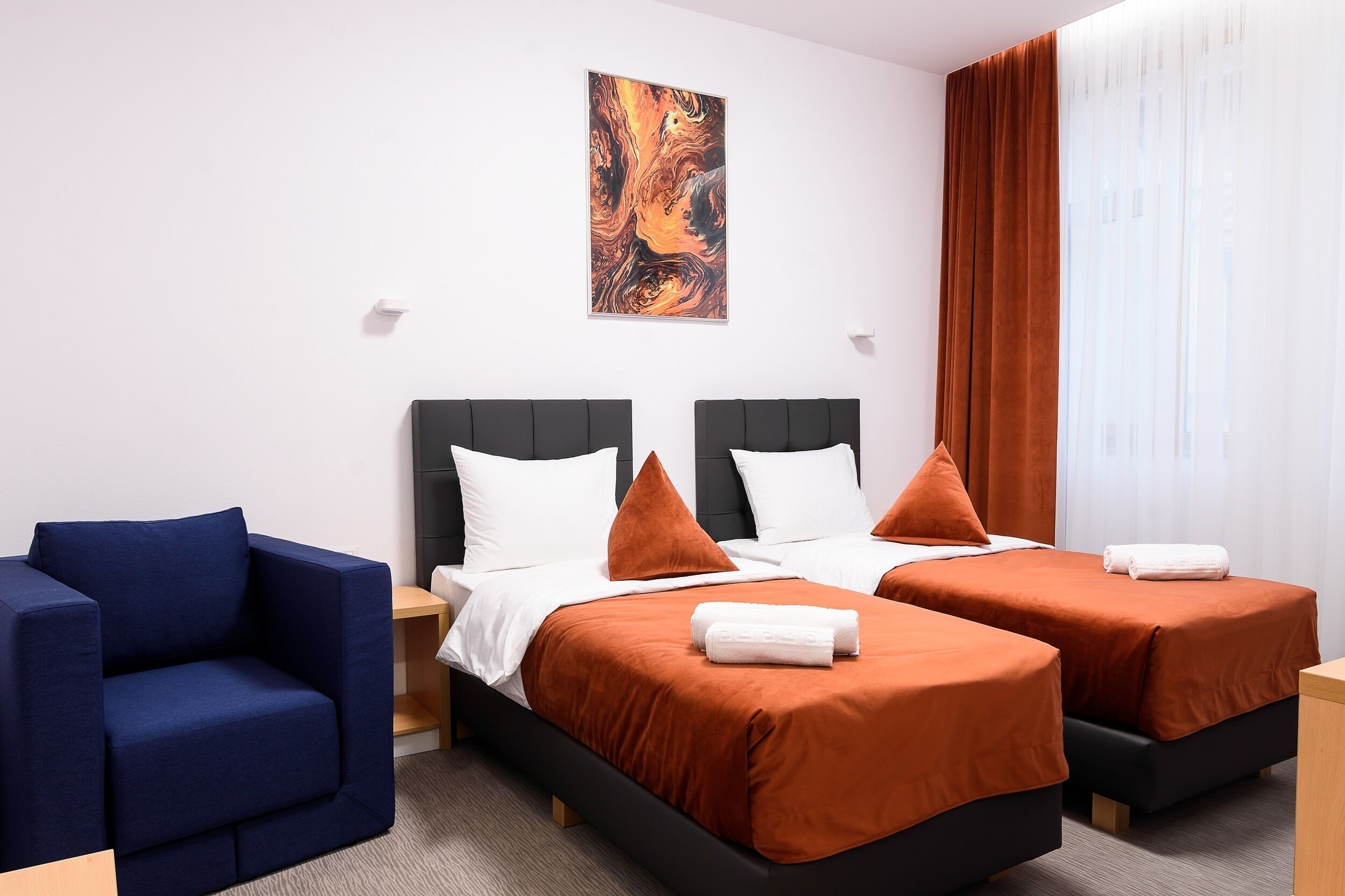 Quarto de hotel com duas camas, uma poltrona azul e um quadro na parede.