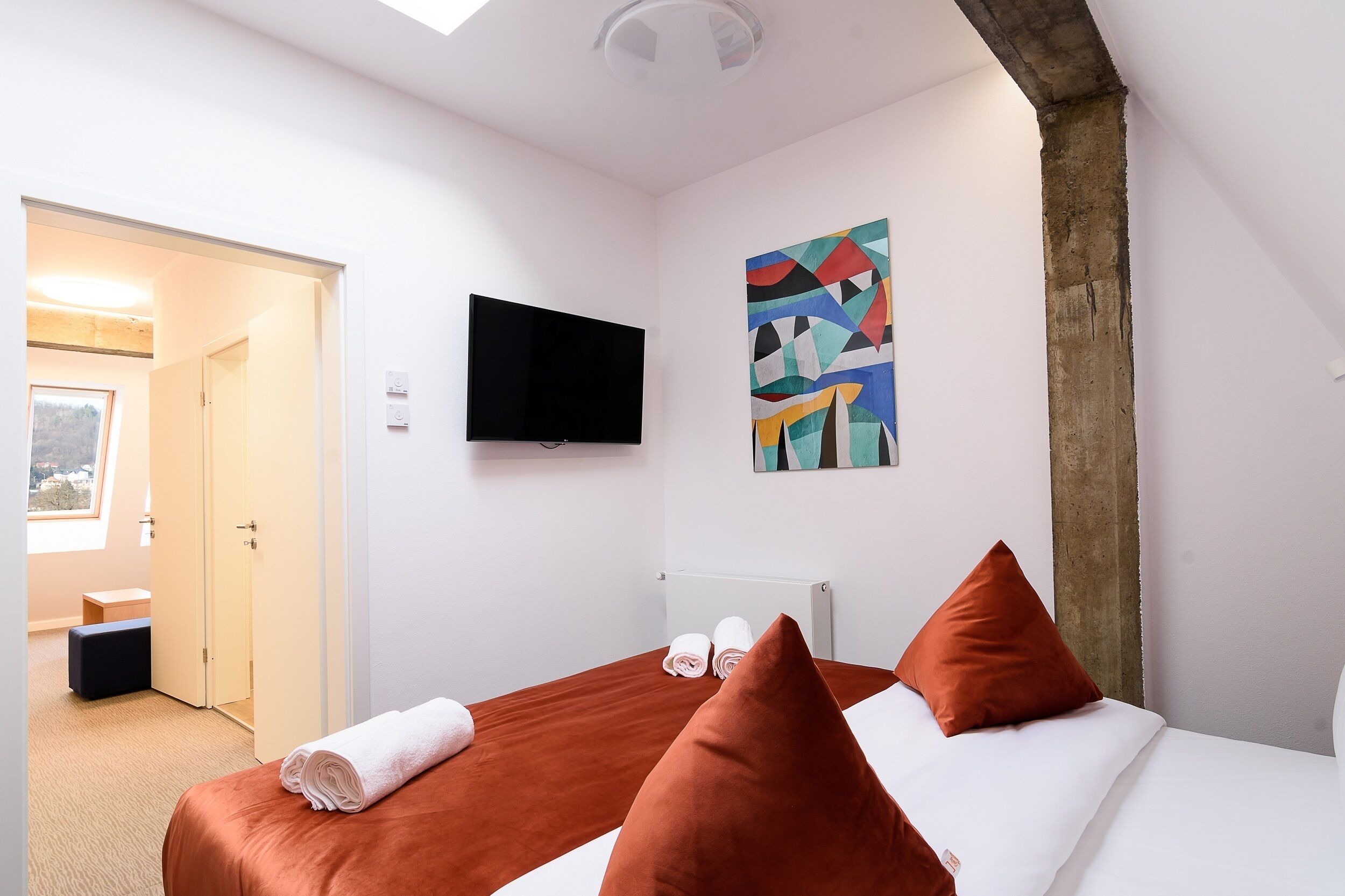 A hotel room with a bed, a TV, a painting, and a window.