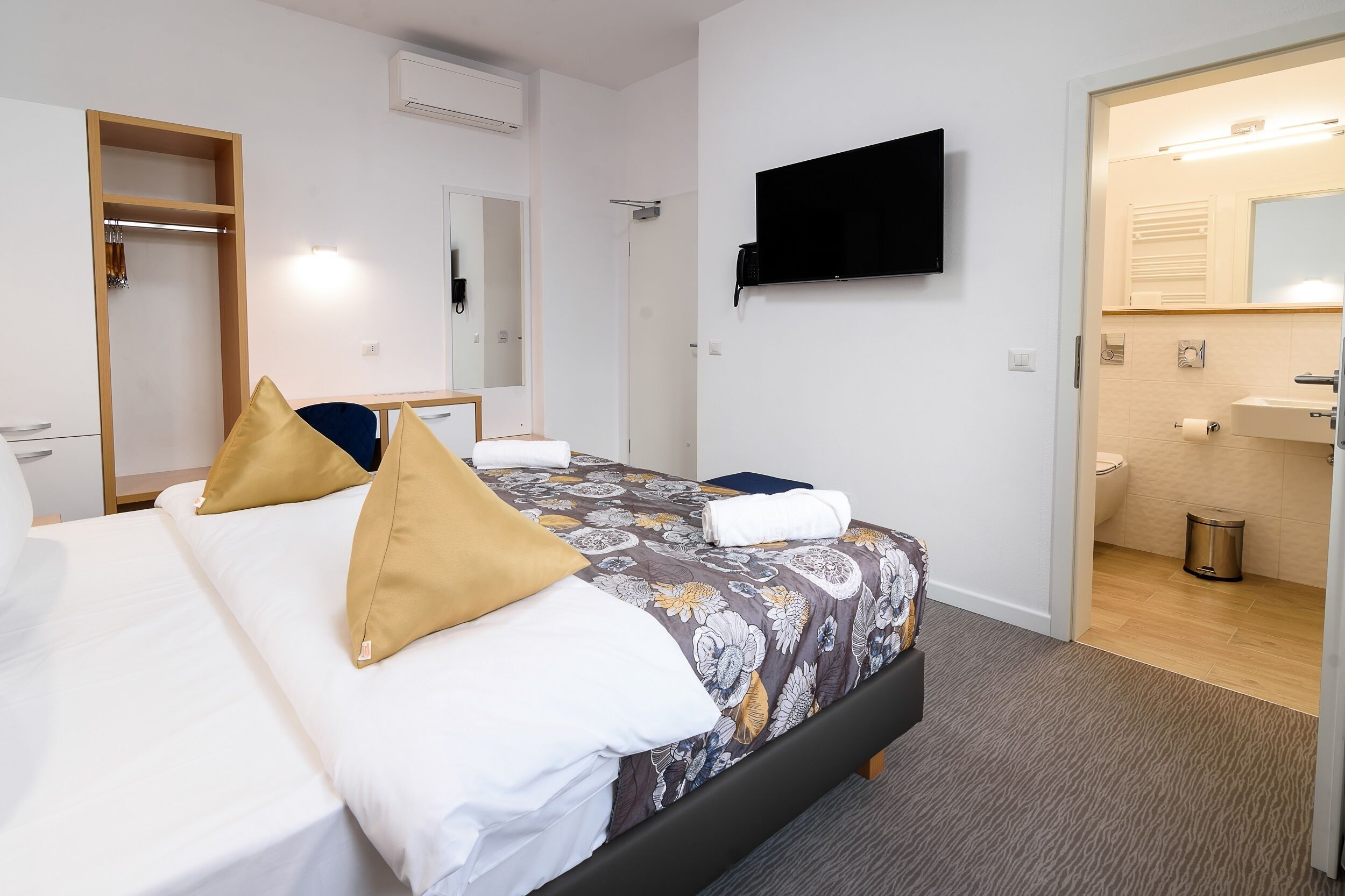 A hotel room with a bed, a TV, a mirror, and a bathroom visible through an open door.