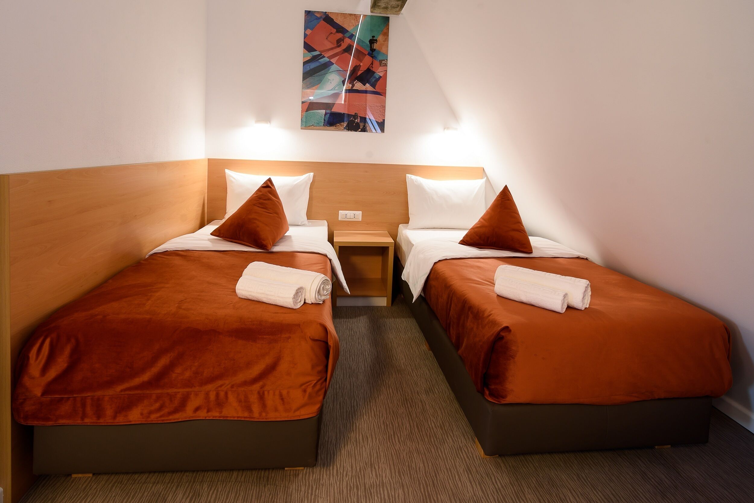 A hotel room with two beds, each with a brown headboard, white pillows, and a white towel. There is a small wooden nightstand between the beds, and a colorful abstract painting on the wall above the beds.