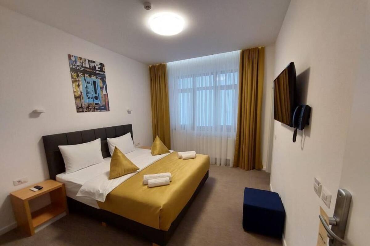 A hotel room with a bed, a bedside table, a TV mounted on the wall, and a window with curtains.
