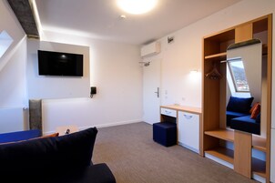 A modern room with a flat-screen TV mounted on the wall, a wooden wardrobe with a mirror, a desk with drawers, and a blue sofa.