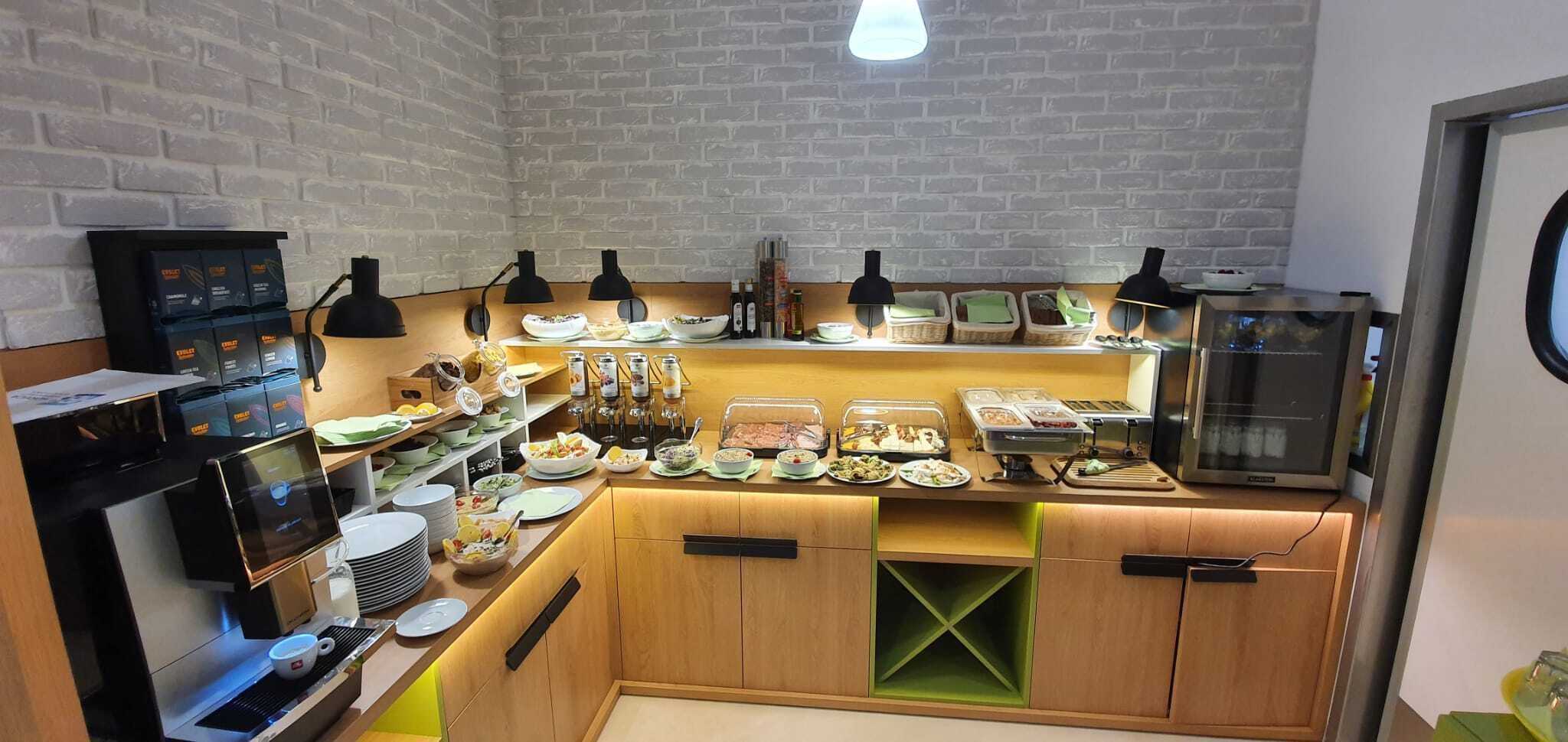 A buffet setup with various food items and condiments, a coffee machine, and a refrigerator.