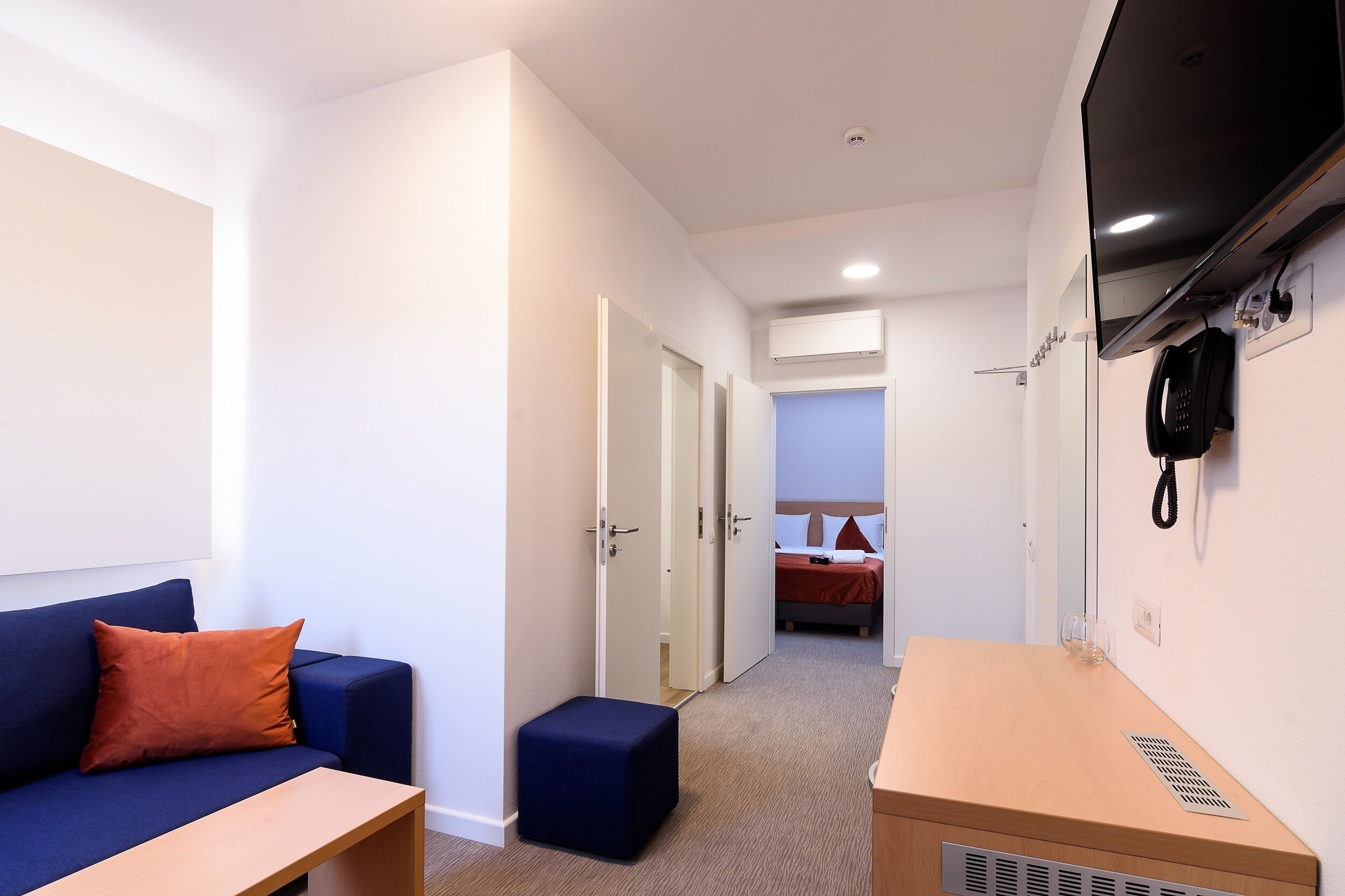Habitación de hotel con un sofá azul, una mesita de madera, un otoman azul, cama con ropa de cama roja y un teléfono de pared.