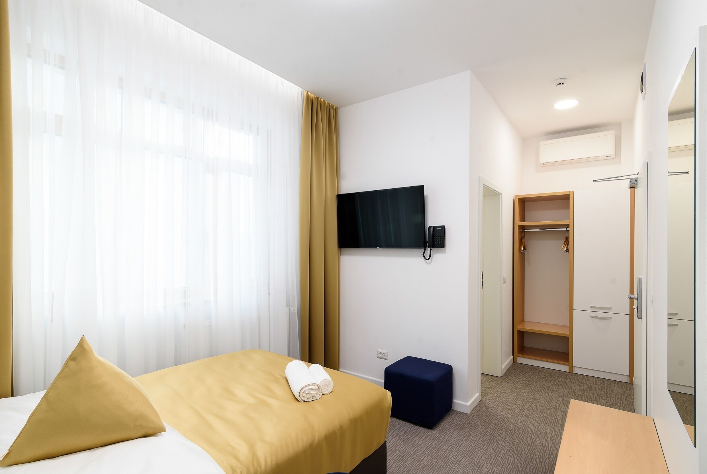A hotel room with a bed, a TV mounted on the wall, a wardrobe, and a small table.