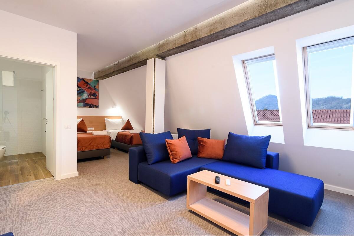 A modern living room with a blue sofa, orange cushions, a wooden coffee table, and a large window with a view of a red-roofed building.
