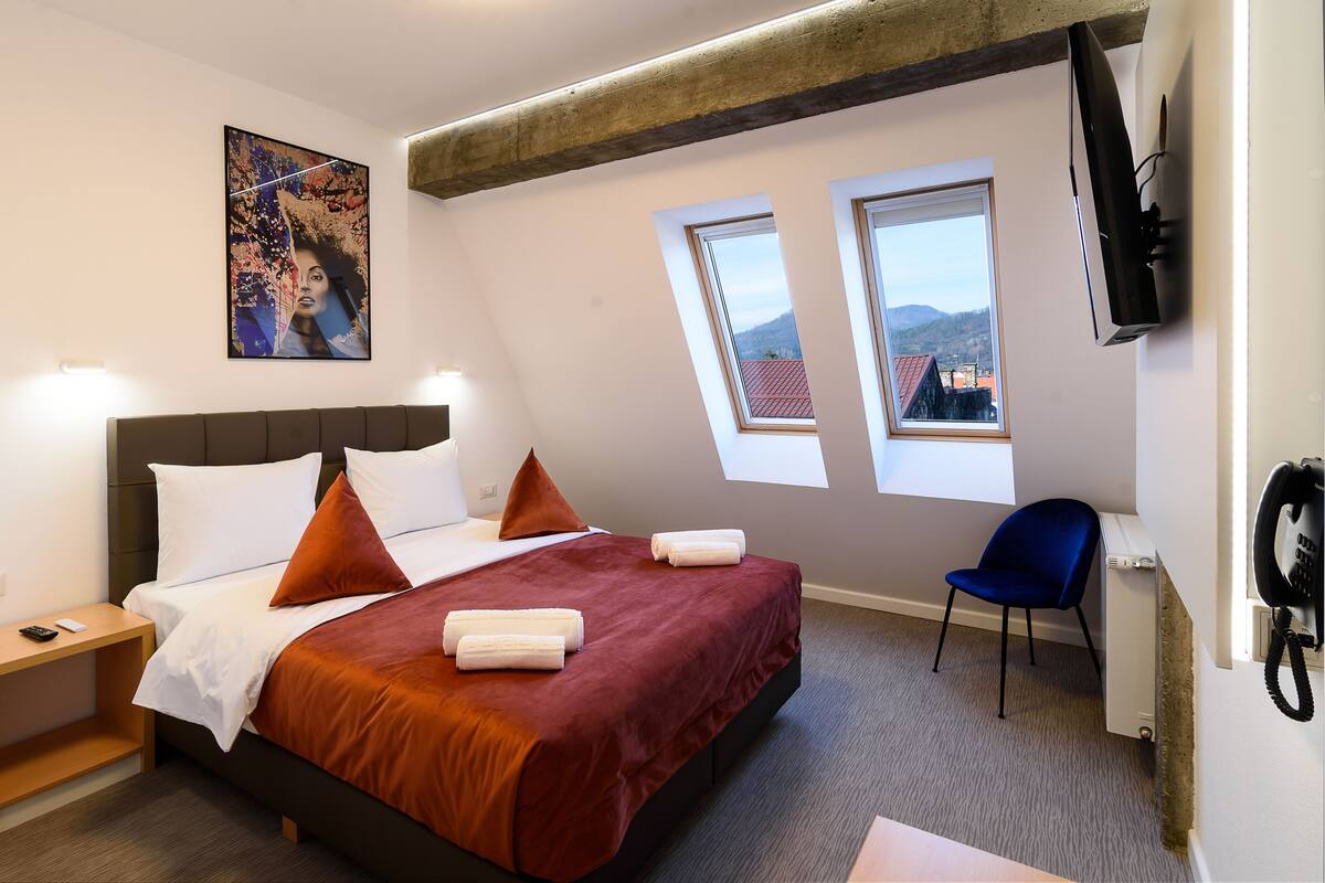 A hotel room with a bed, two windows, a TV, and a chair.