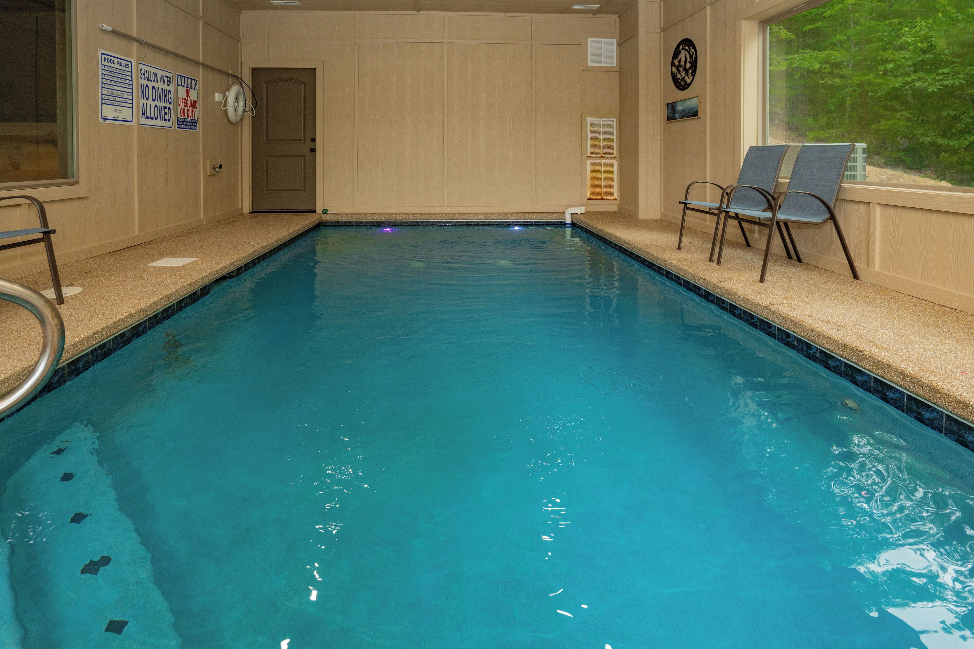 An indoor swimming pool with a clear water view, a pool ladder, and two lounge chairs by the window.