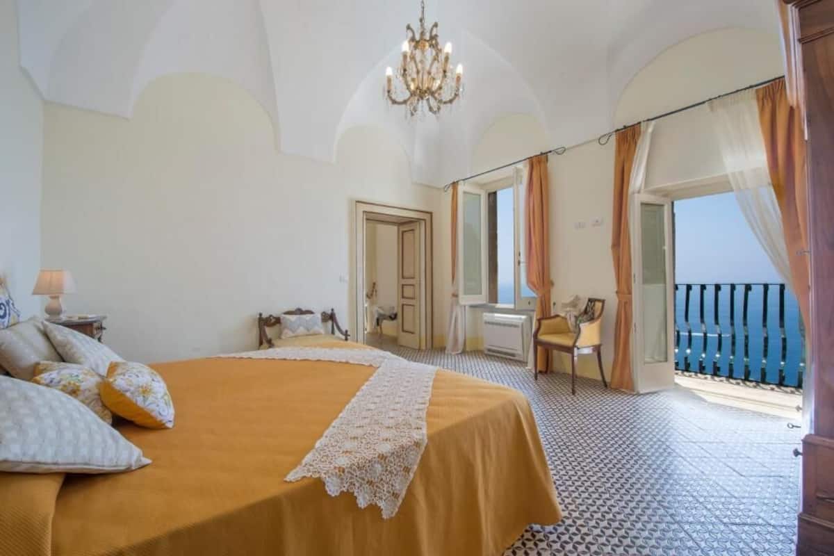 A bedroom with a large bed, a chandelier, a view of the sea, and a balcony with a chair.