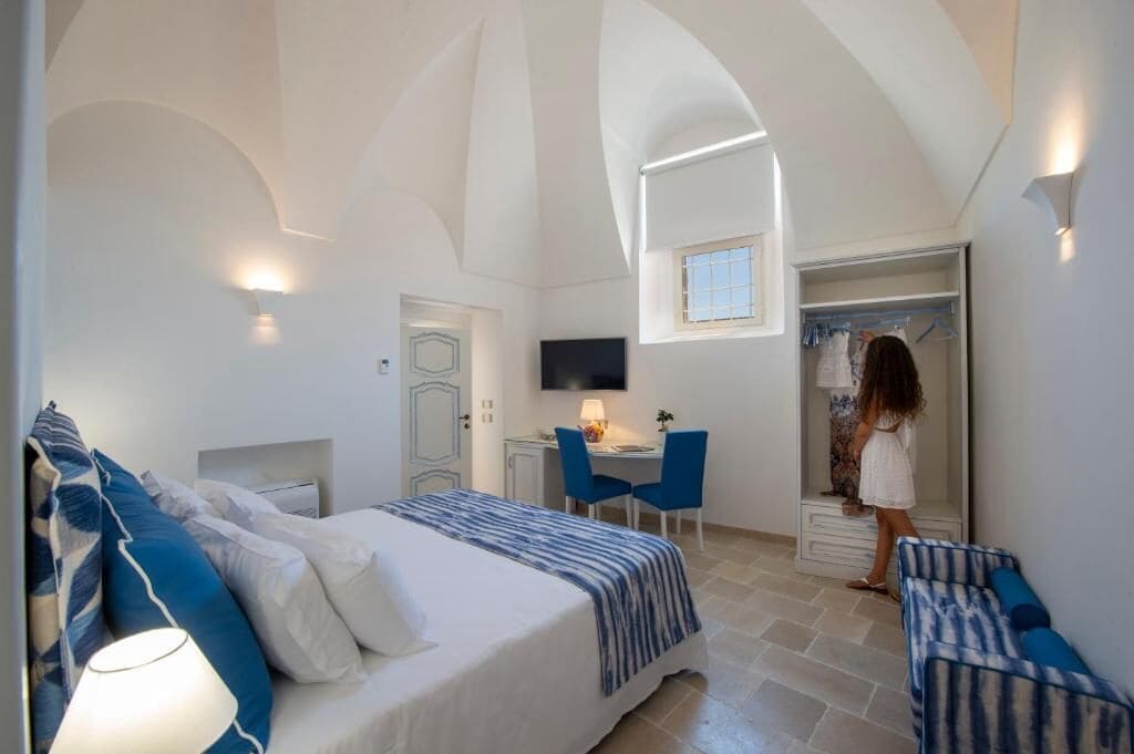 A bedroom with a bed, a desk, and a chair. There is a TV on the wall and a window.