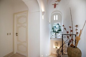 Un pasillo con una puerta blanca, una escalera con barandilla y un jarrón decorativo con ramas.
