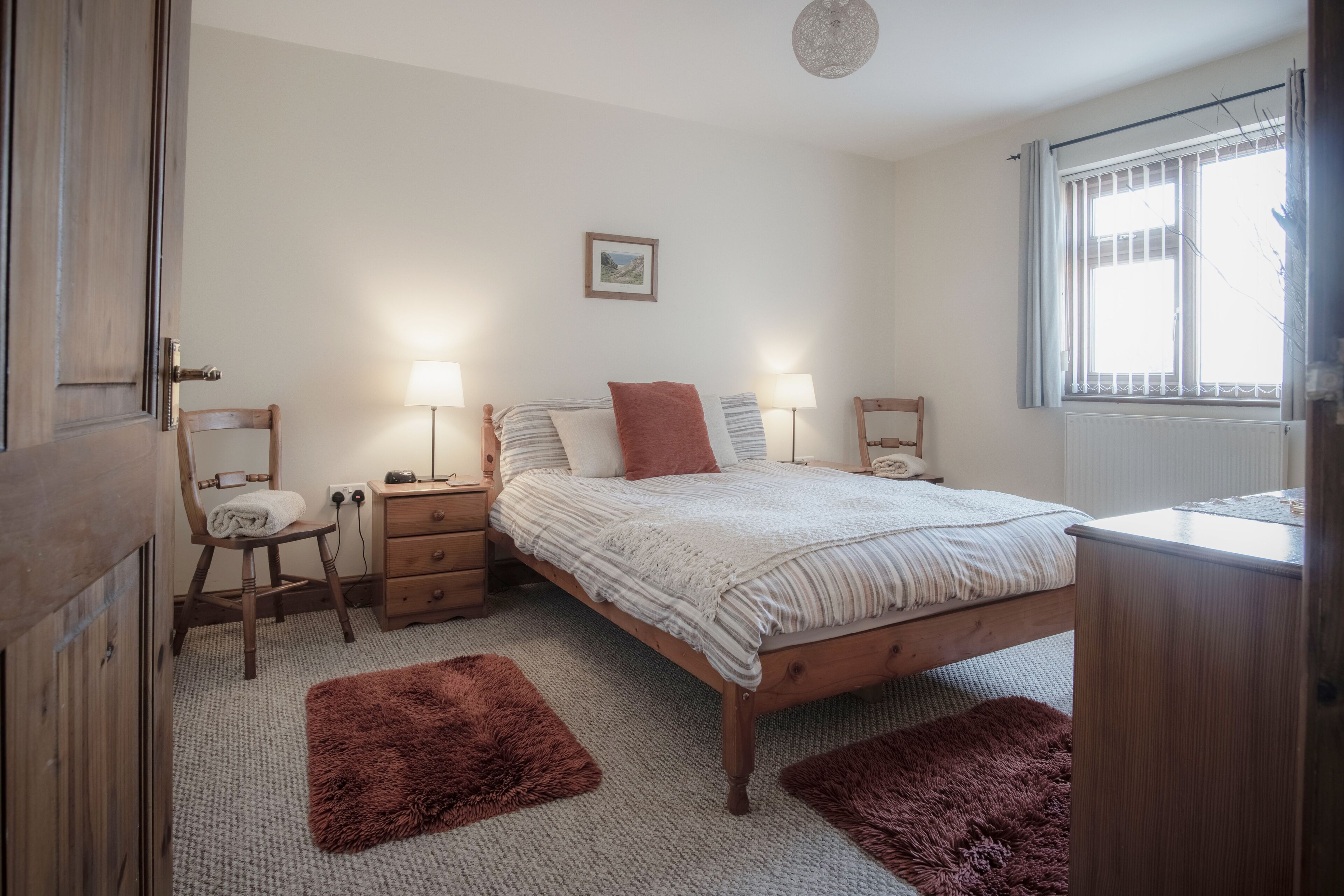 Un dormitorio con una cama de madera, dos mesitas de noche, una silla, una lámpara y una ventana con barrotes.