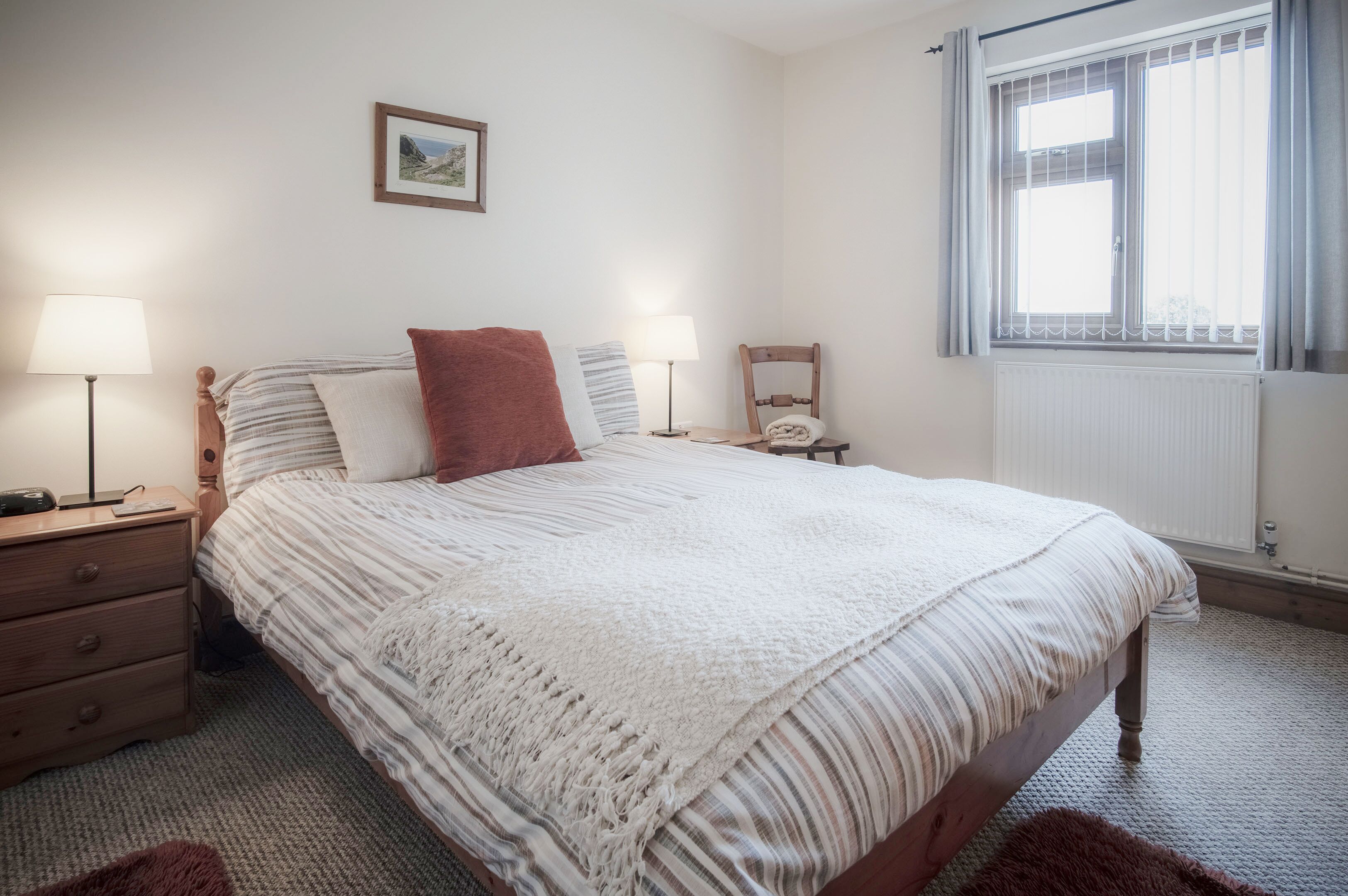 Cama bem arrumada com colcha branca e listrada, duas luminárias de cabeceira, mesa de cabeceira de madeira, uma cadeira e janela com persianas.