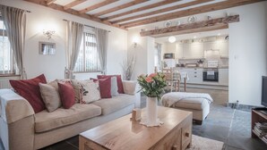 Een gezellige woonkamer met een bank, salontafel en een bloevas. In de achtergrond is een keuken zichtbaar met witte kasten en een eethoek.