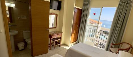 A hotel room with two beds, a desk, a TV, and a balcony with a view of the sea.