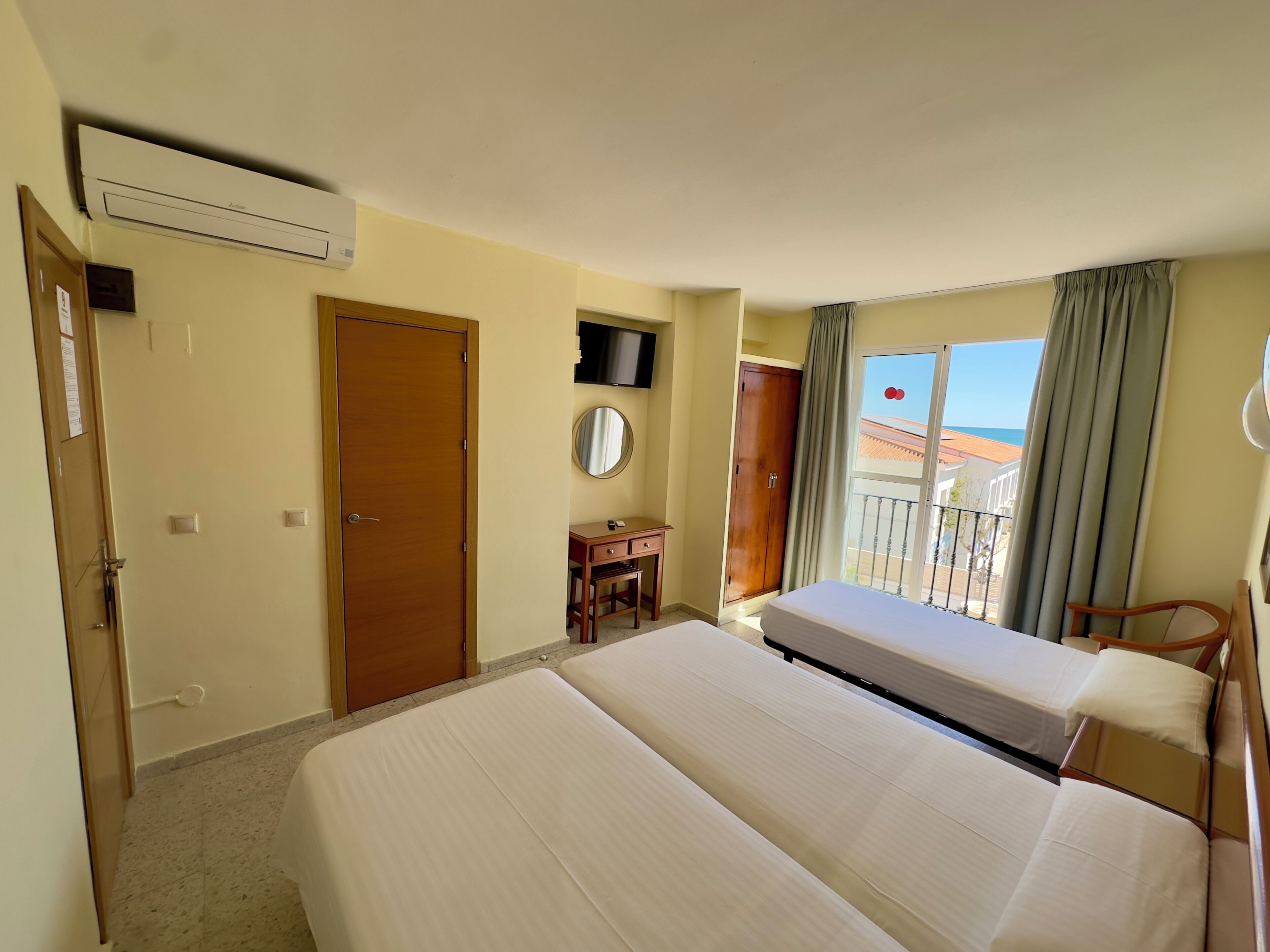 A hotel room with two beds, a wooden wardrobe, a small desk, a TV, and a balcony with curtains.