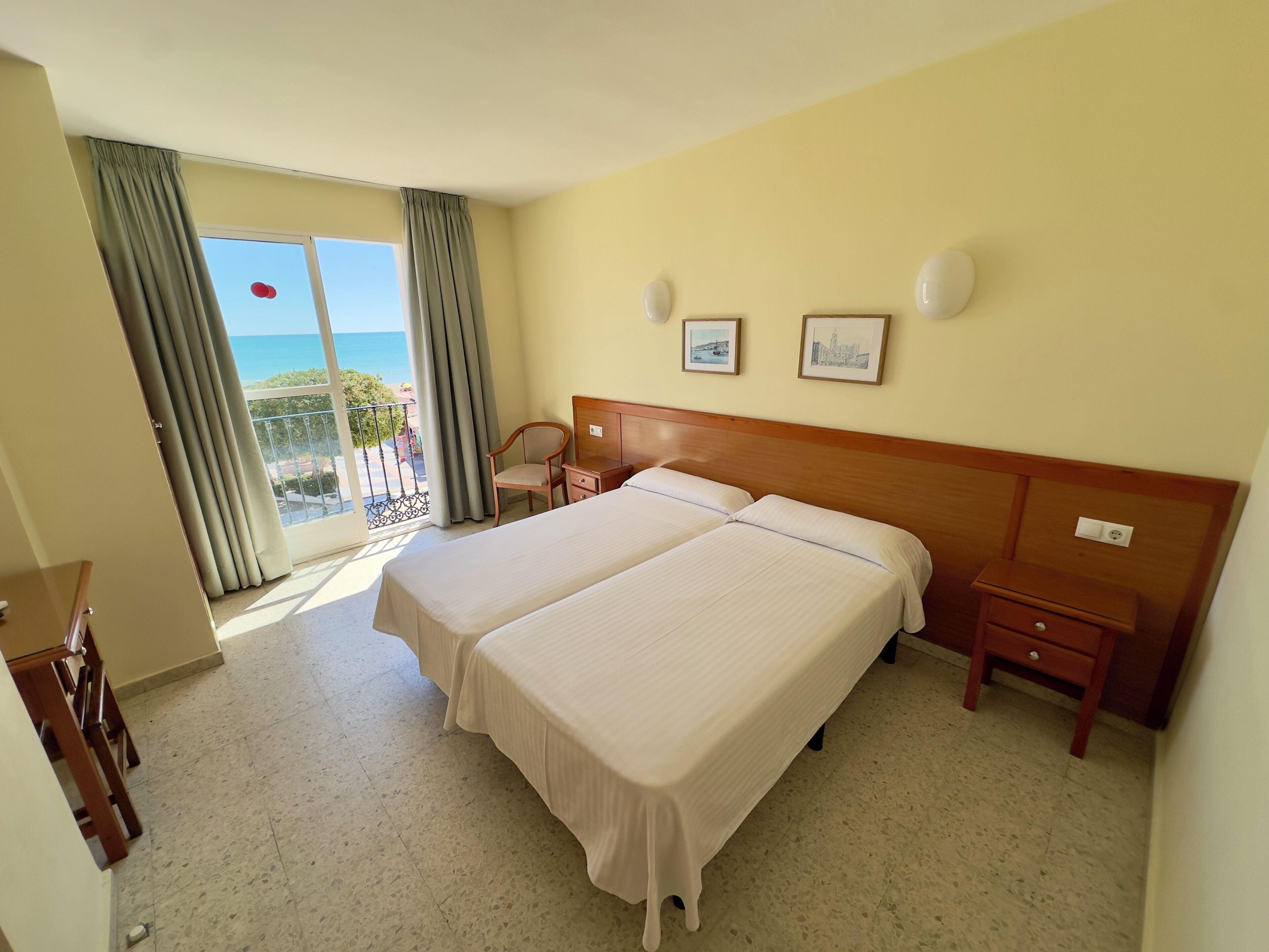 A hotel room with two beds, a chair, a nightstand, a window with curtains, and a balcony with a view.