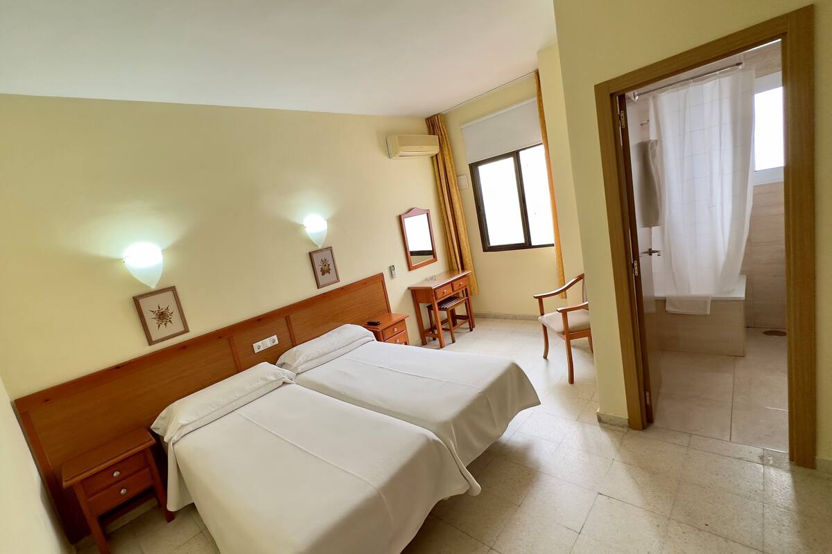 A hotel room with two beds, a desk, a chair, a window with curtains, and a door.