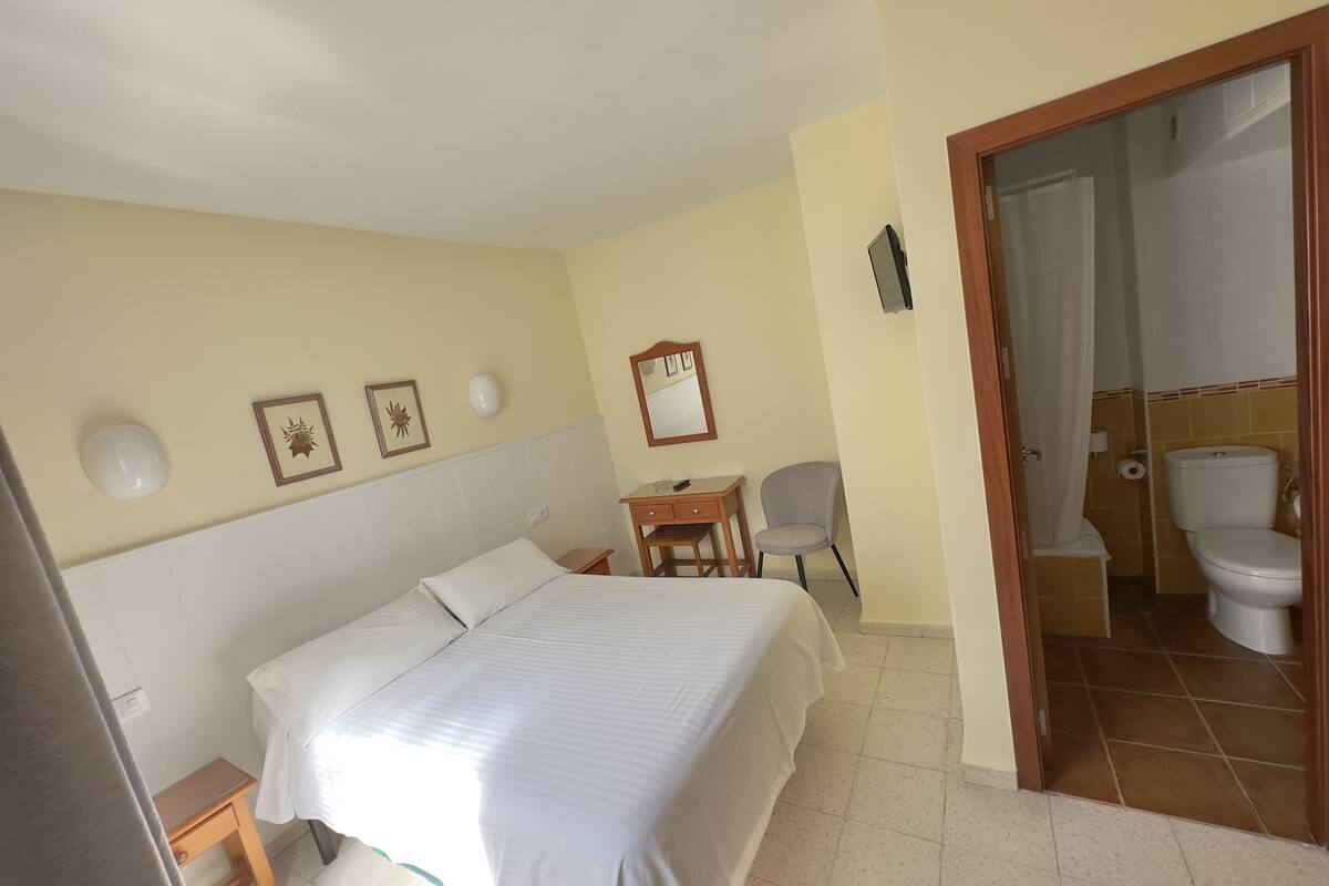 A hotel room with a bed, a desk, a chair, a bathroom with a toilet and sink, and a door leading to another room.