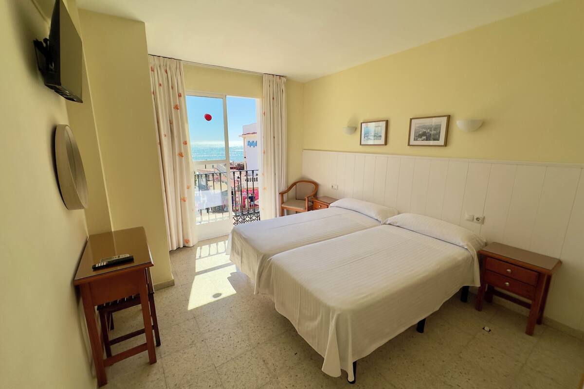 A hotel room with two beds, a TV, a small table, and a balcony with a view.