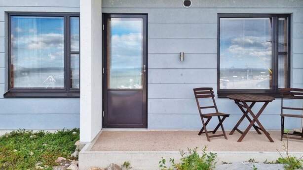 Una casa moderna con una puerta oscura, dos ventanas con persianas, un pequeño patio con una mesa y sillas de madera, y un cimiento de hormigón.