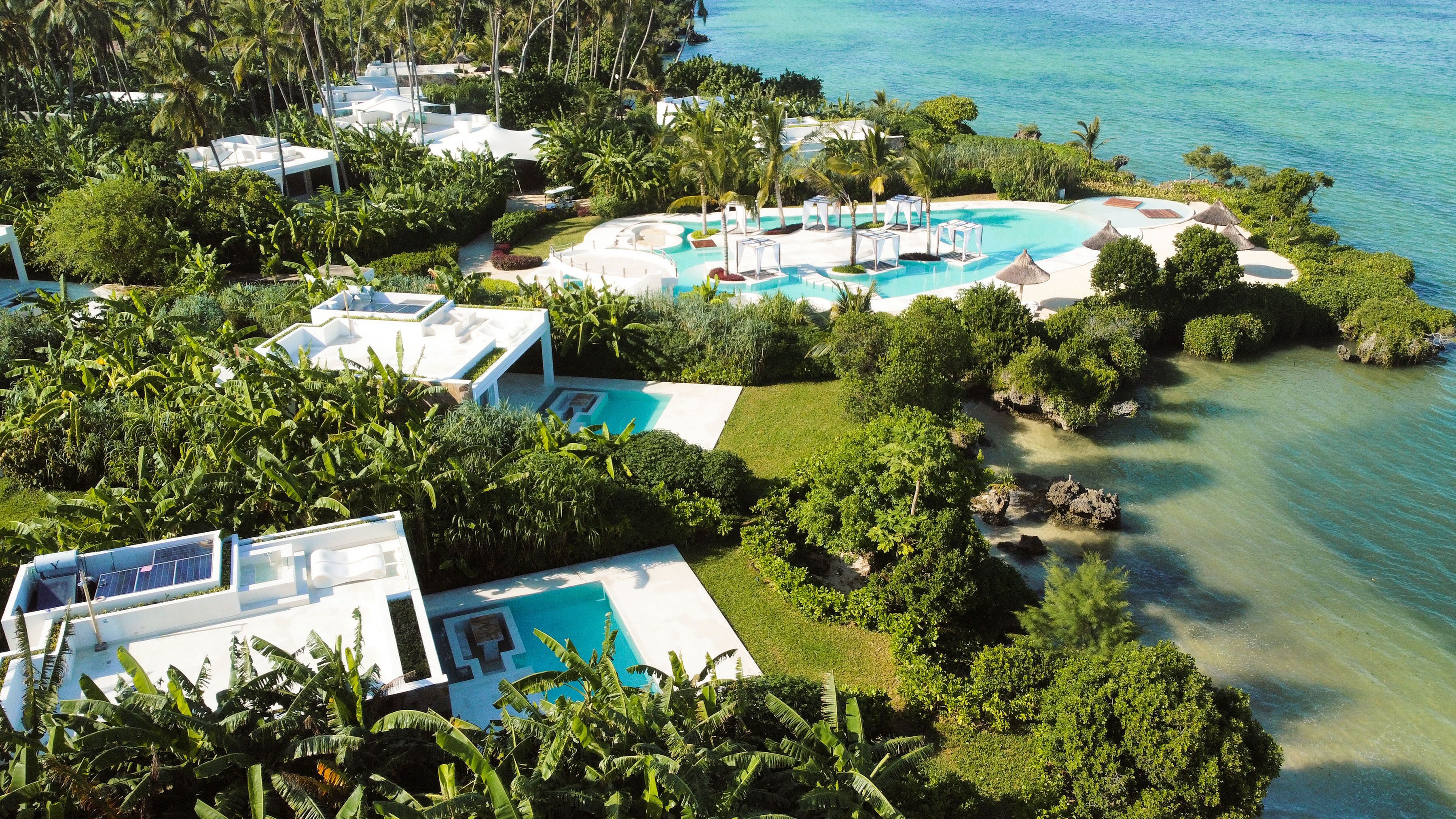 An aerial view of a resort with multiple swimming pools, lush greenery, and a clear view of the ocean.
