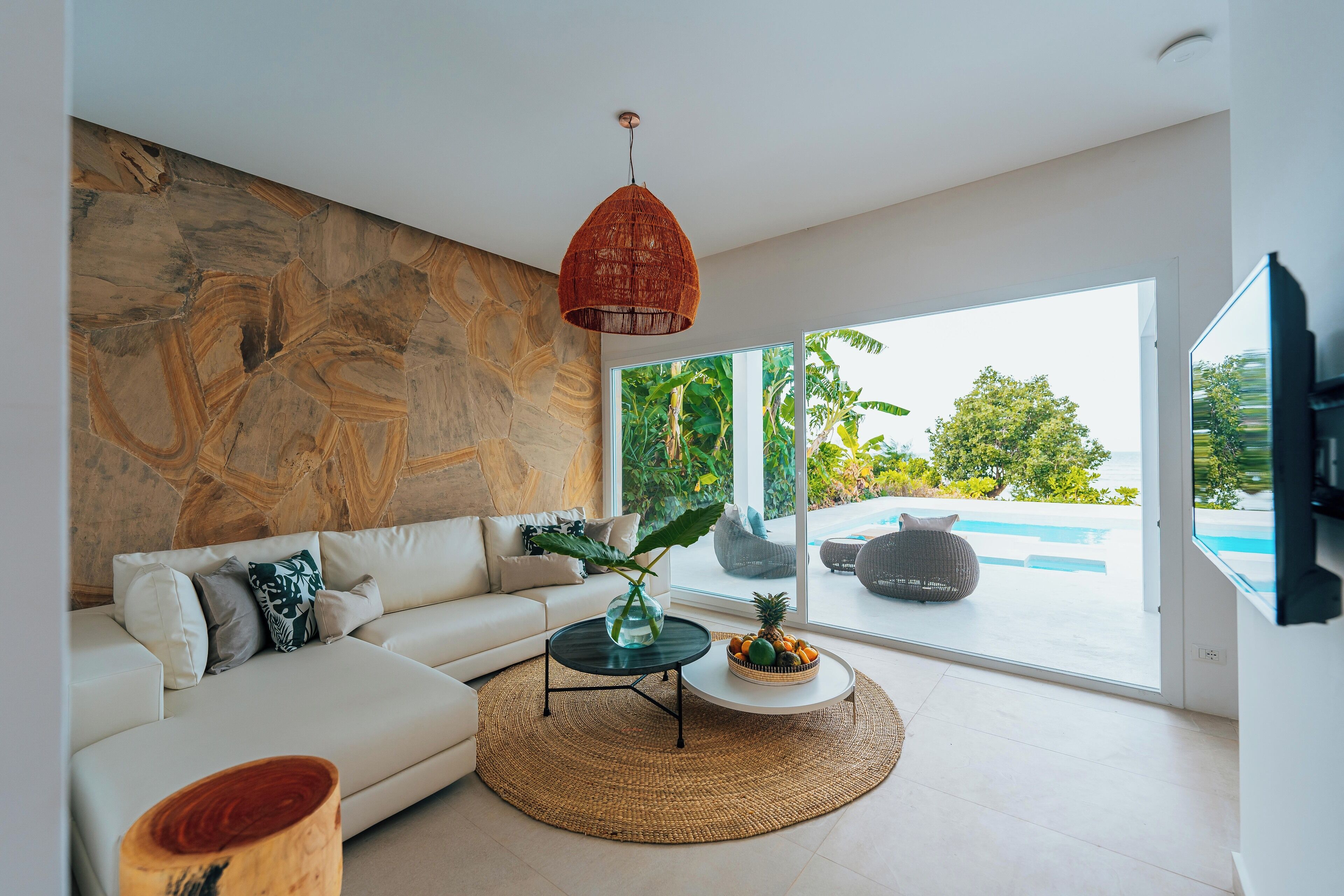 A modern living room with a white sofa, a round coffee table, and a large window offering a view of a swimming pool and greenery.
