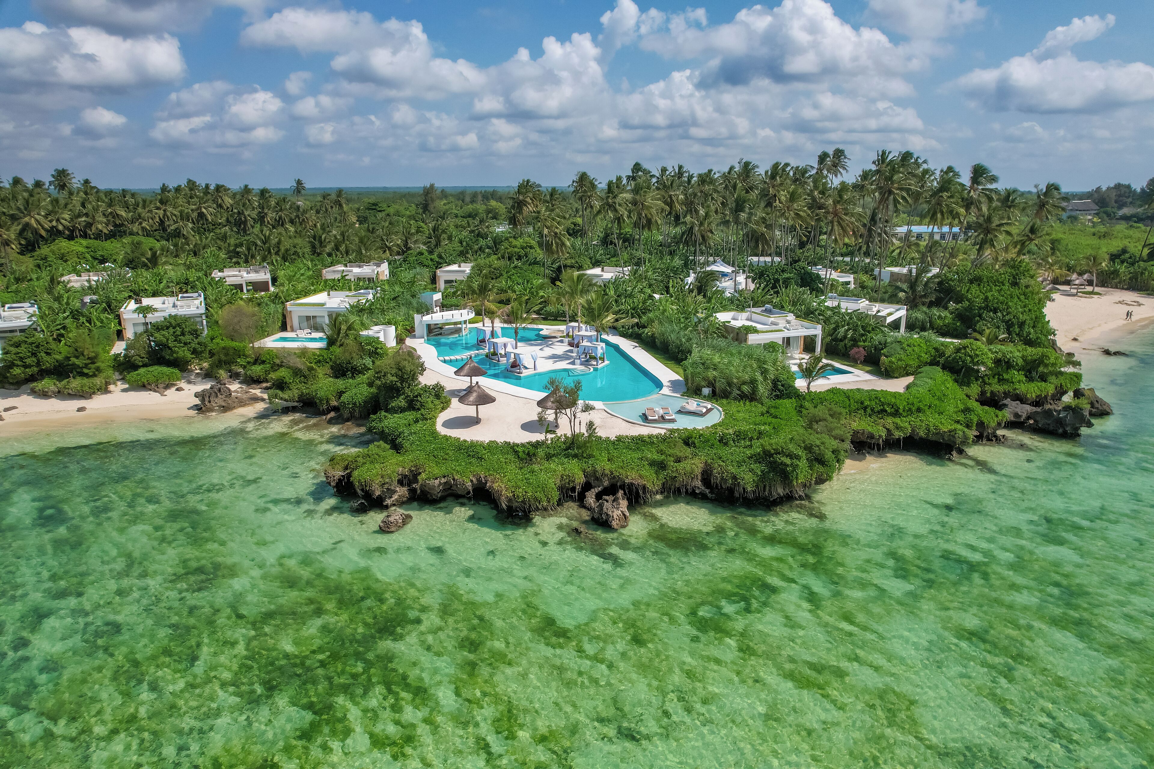 Vista aérea de un complejo turístico con una piscina central, rodeado de exuberante vegetación y aguas turquesas cristalinas.