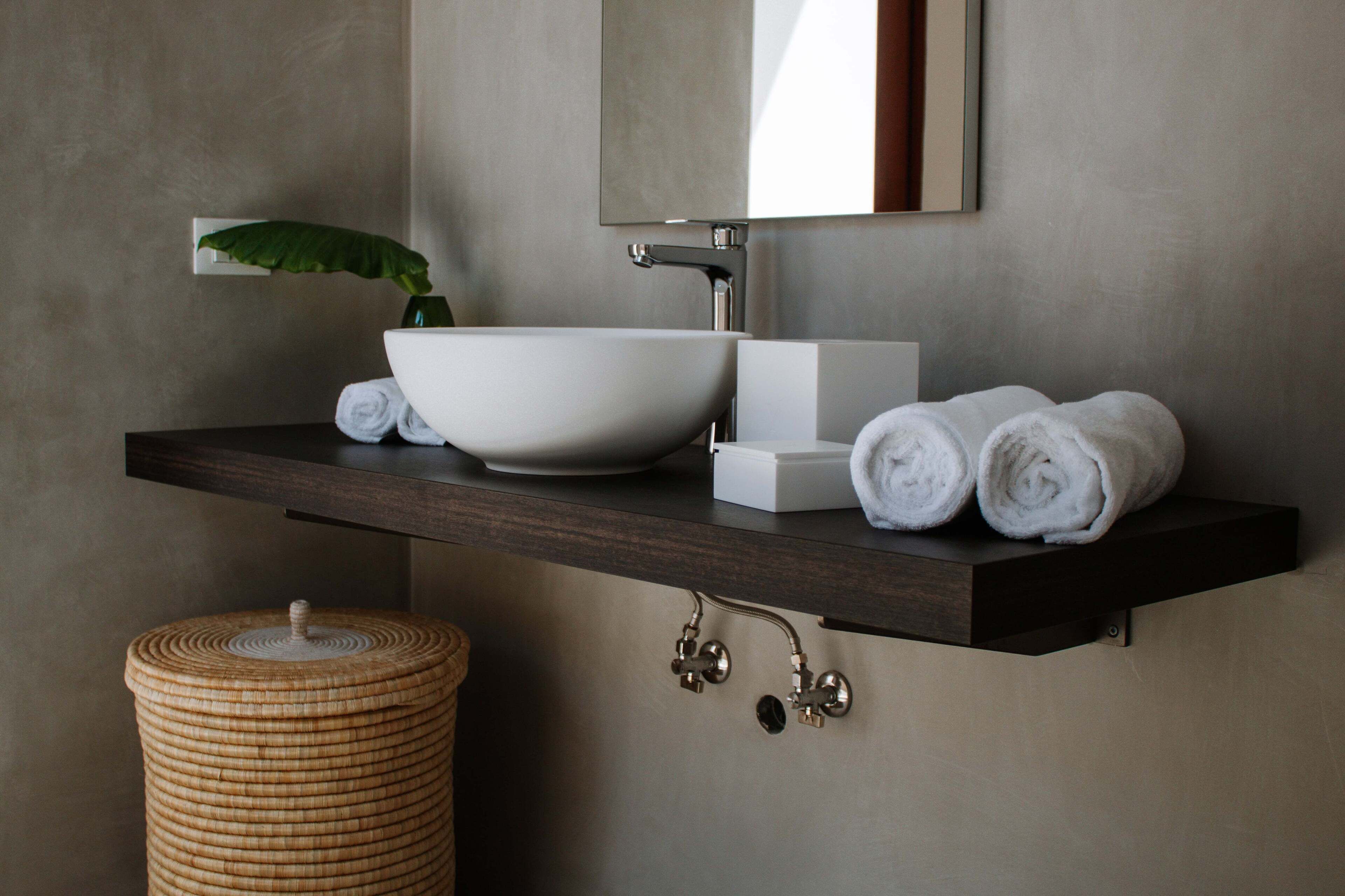 Un baño moderno con un lavamanos blanco de diseño, un estante de madera oscura con toallas enrolladas, una pequeña planta en maceta y un cesto de mimbre.