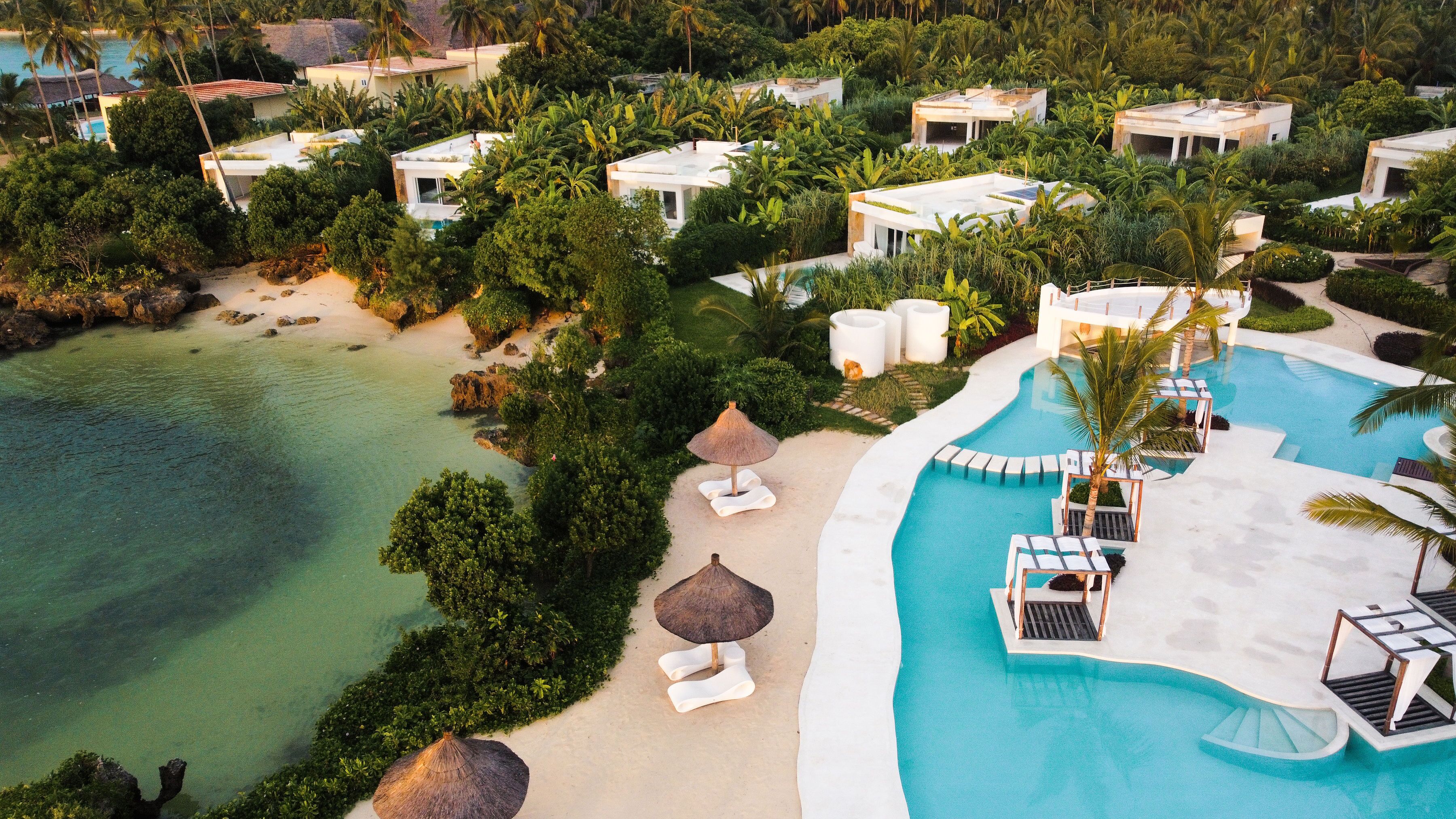 An aerial view of a resort with multiple white buildings surrounded by lush greenery and a clear blue pool.