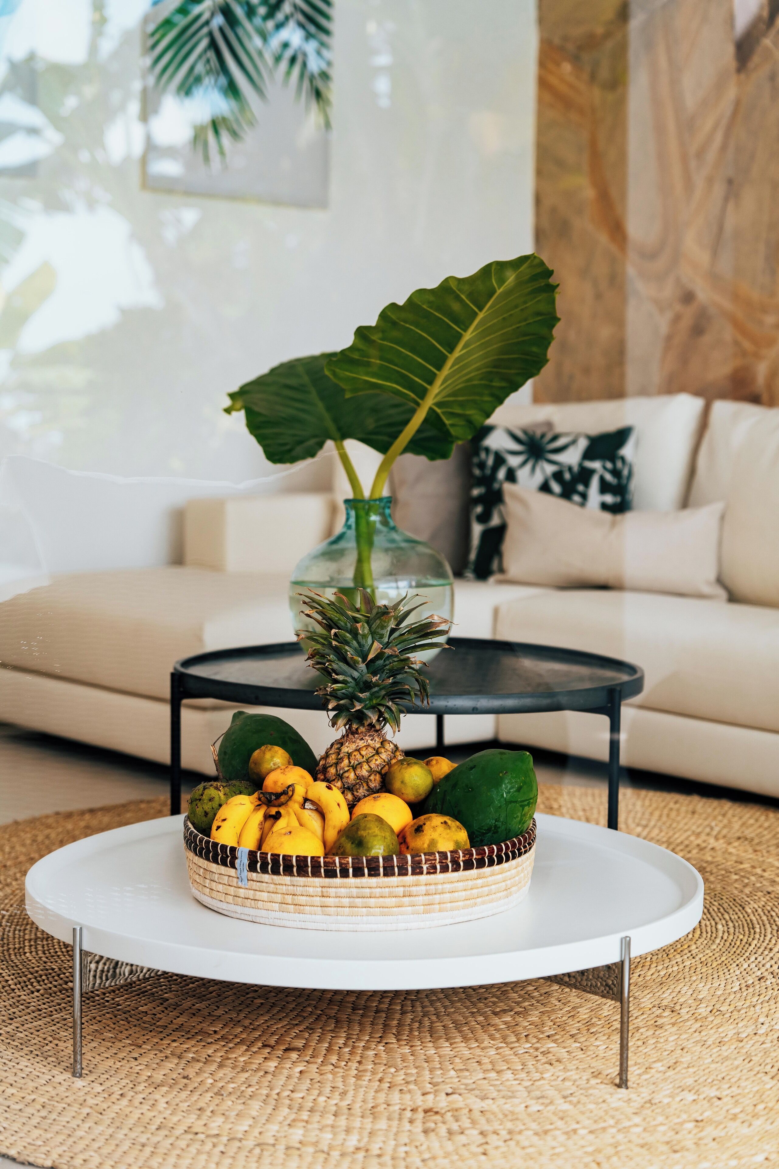 A round white table with a woven basket of fruits and a glass vase with a plant.