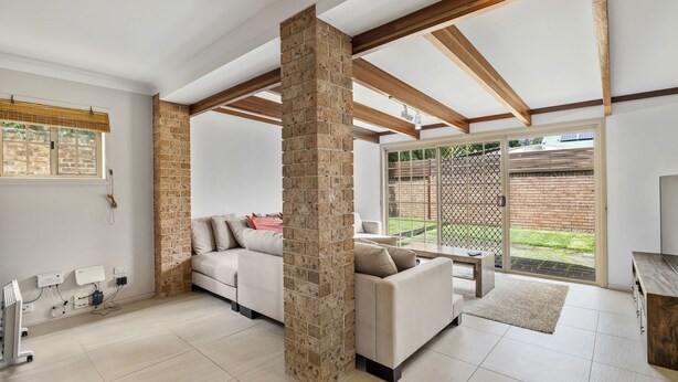 A spacious living room with a high ceiling, exposed wooden beams, a brick column, and a sliding glass door leading to a backyard. - Palm Beach House - Red Rock location (Red Rock)