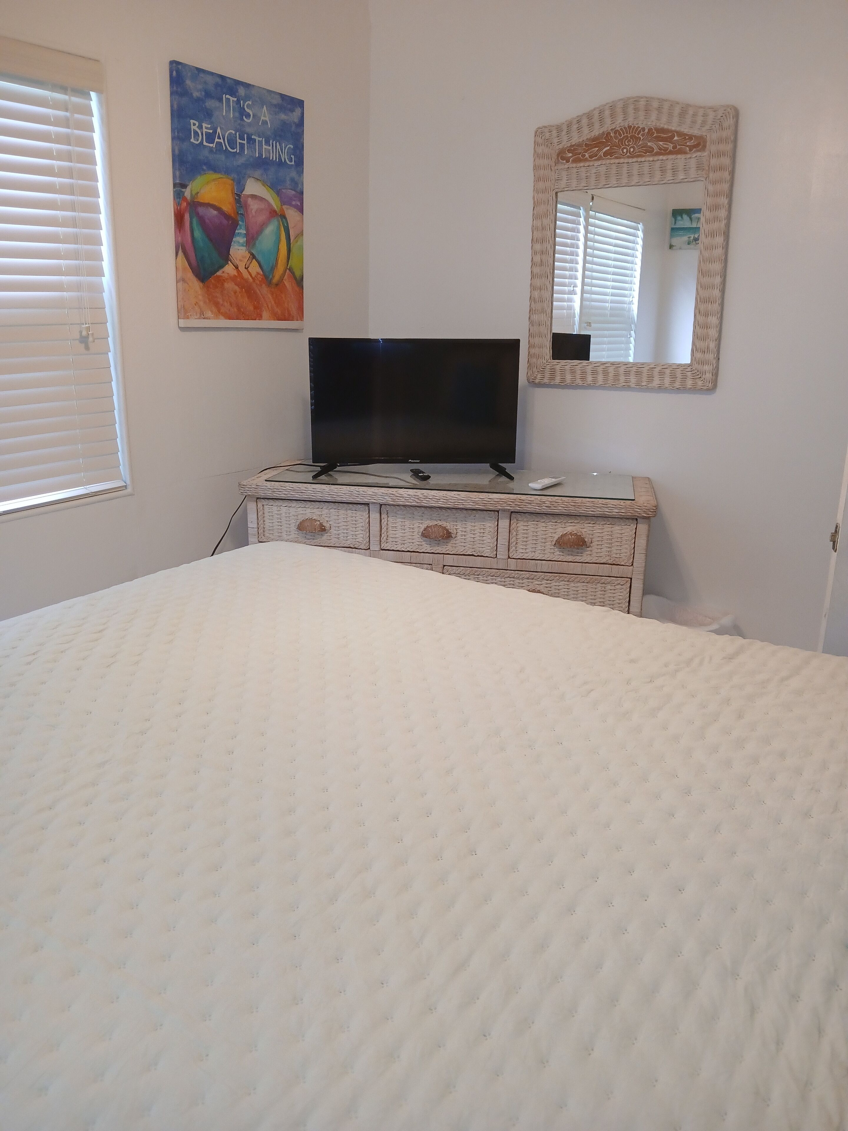 A bedroom with a bed, a television on a dresser, a beach-themed painting, and a mirror.