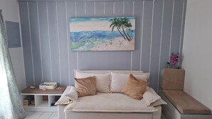 En stue med en beige sofa, et maleri af en strandscene, et lille sofabord og en bænk.
