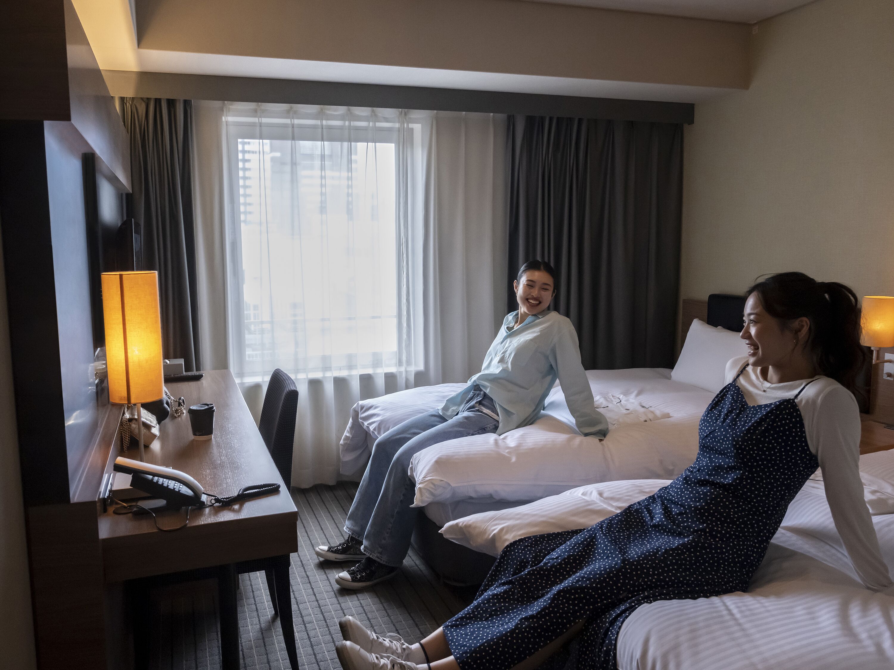 Une chambre d’hôtel avec deux lits, un bureau avec une lampe et une fenêtre avec des rideaux.