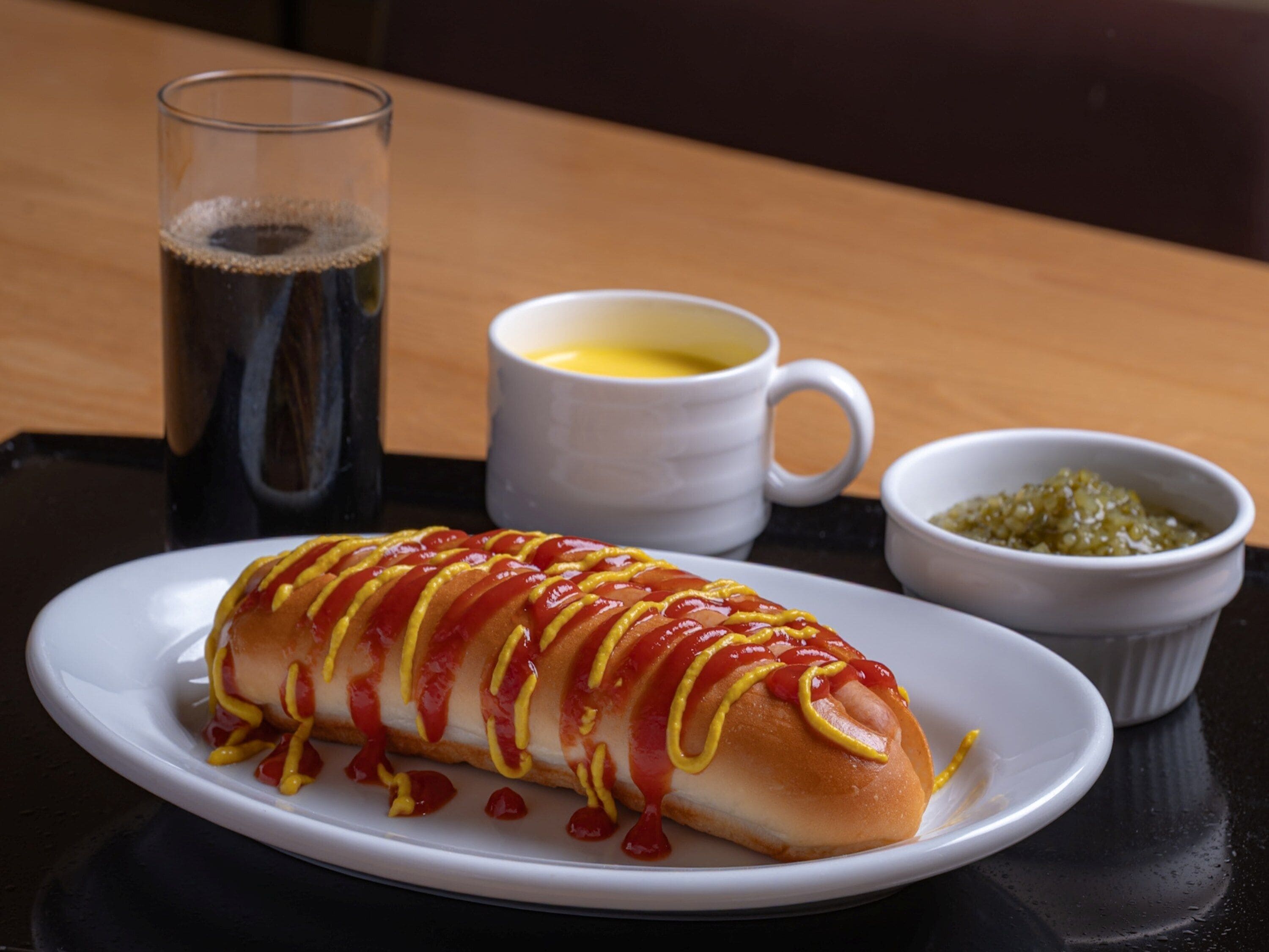 A hot dog with ketchup and mustard, a glass of soda, a cup of coffee, and a small bowl of relish on a table.