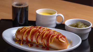 A hot dog with ketchup and mustard, a glass of soda, a cup of coffee, and a small bowl of relish on a table.