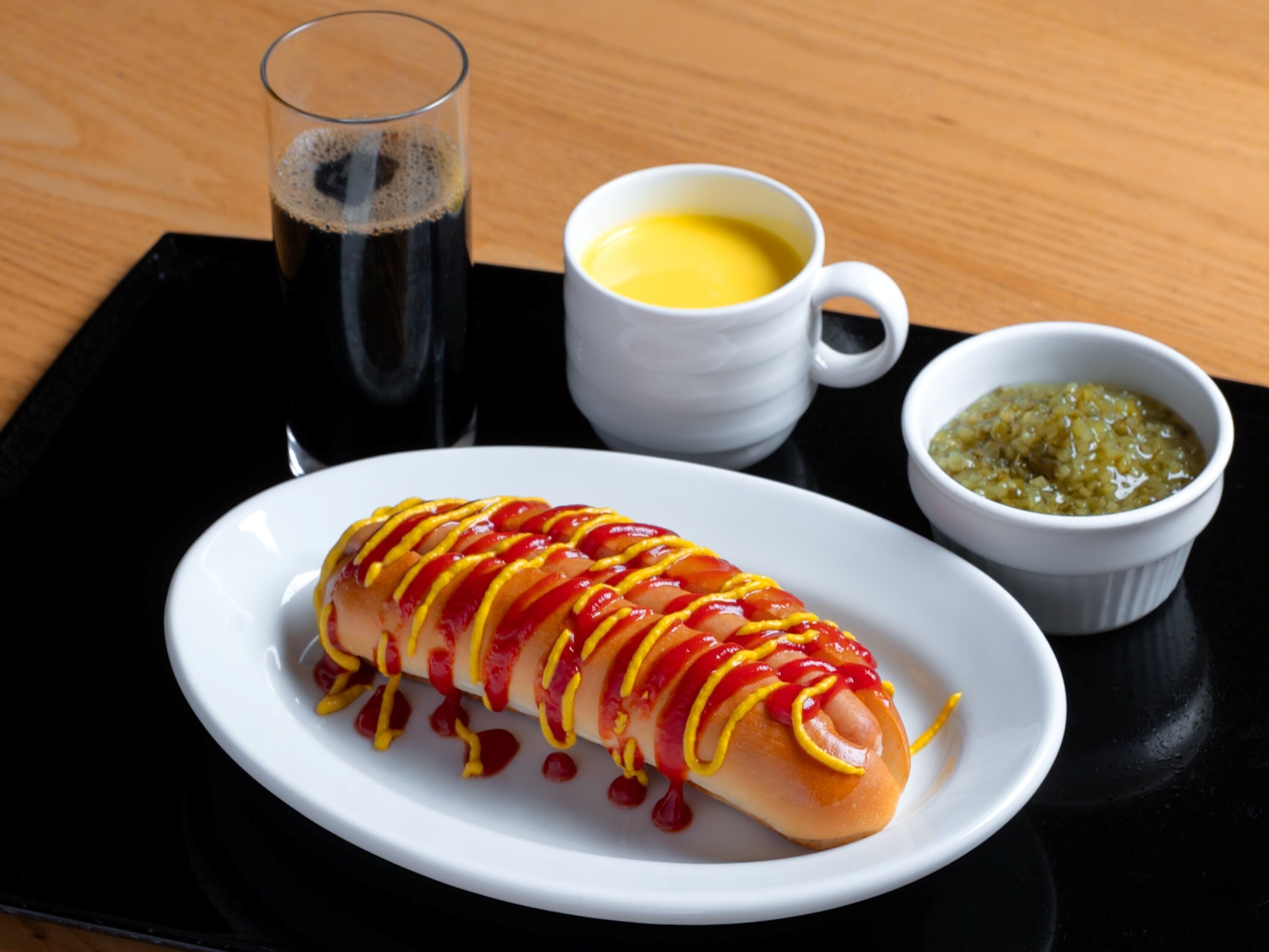 A hot dog with mustard and ketchup, a glass of soda, a cup of coffee, and a small bowl of relish on a black tray.