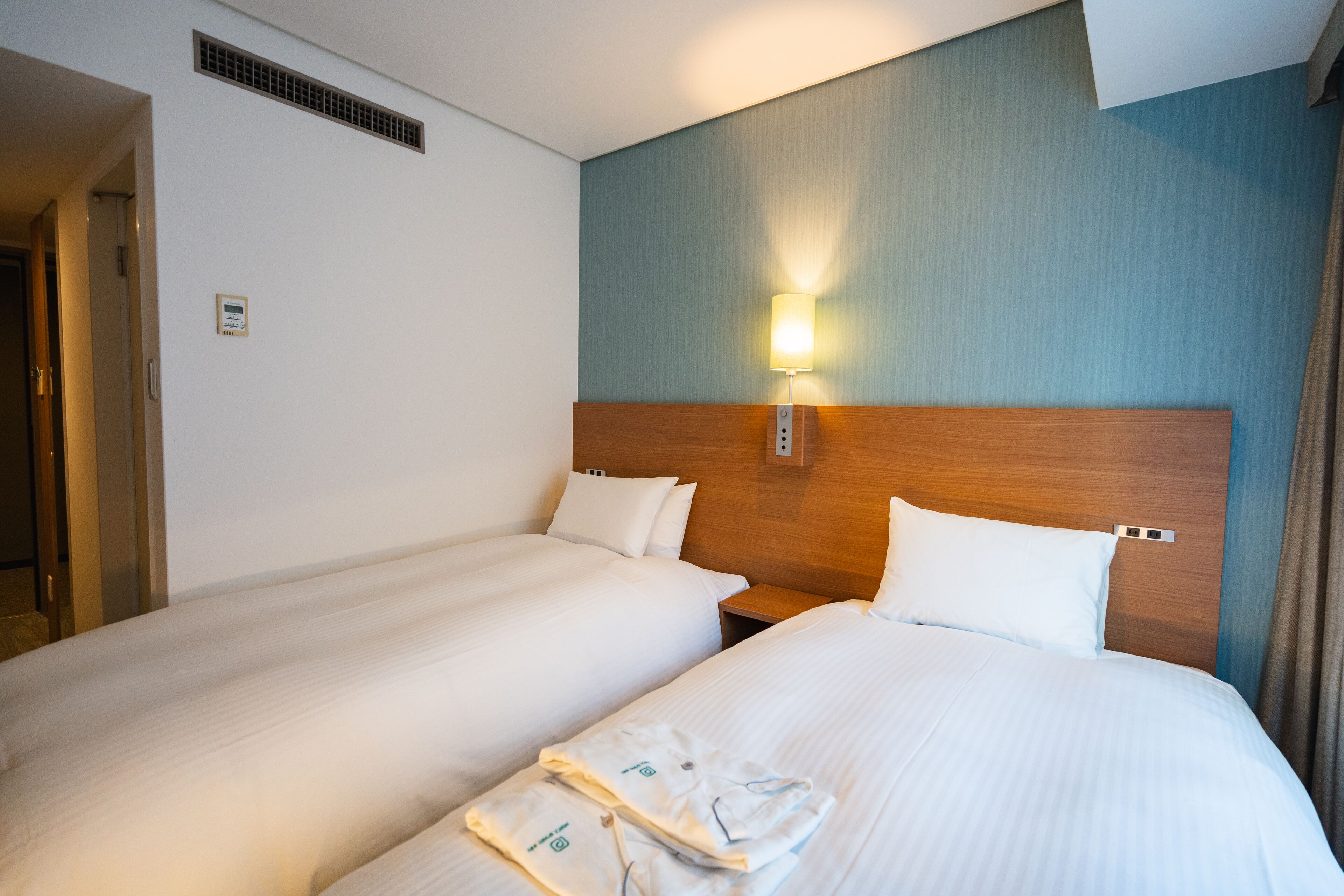 A hotel room with two beds, a wooden headboard, a wall-mounted light, and a visible door.