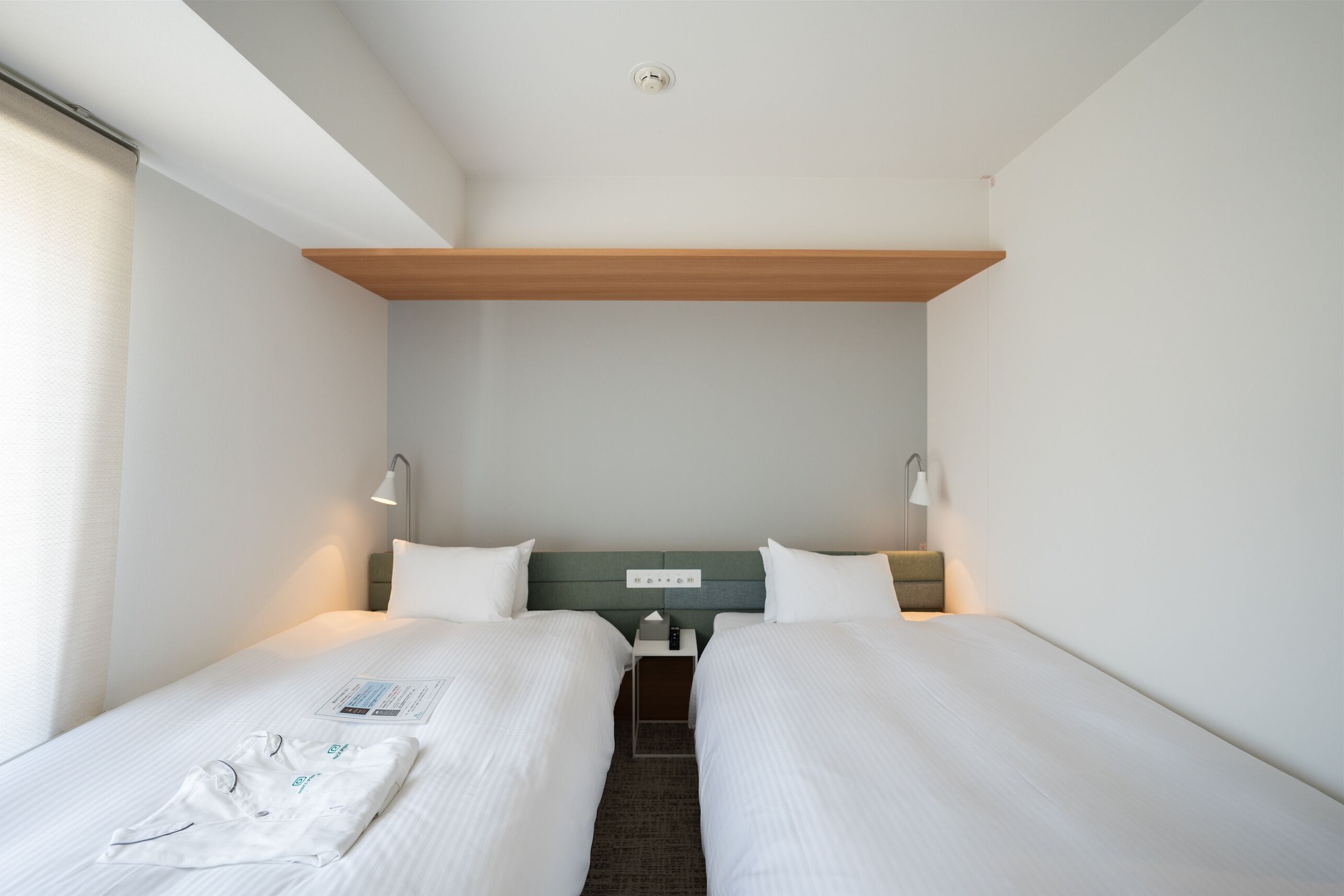 Quarto de hotel com duas camas, uma prateleira de madeira acima, uma mesa de cabeceira com abajur e uma janela com cortinas.