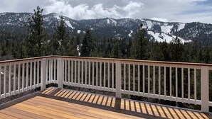 Une terrasse en bois, dotée d’une balustrade blanche, offre une vue sur un paysage montagneux, avec des sommets enneigés et de vastes forêts.