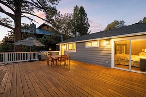 A wooden deck with a grill and outdoor furniture, a glass door leading to a bedroom, and a tree in the background.