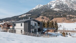 Une maison moderne à deux étages, dotée d’un balcon, entourée d’une clôture en bois, située dans un paysage enneigé avec des montagnes en arrière-plan.