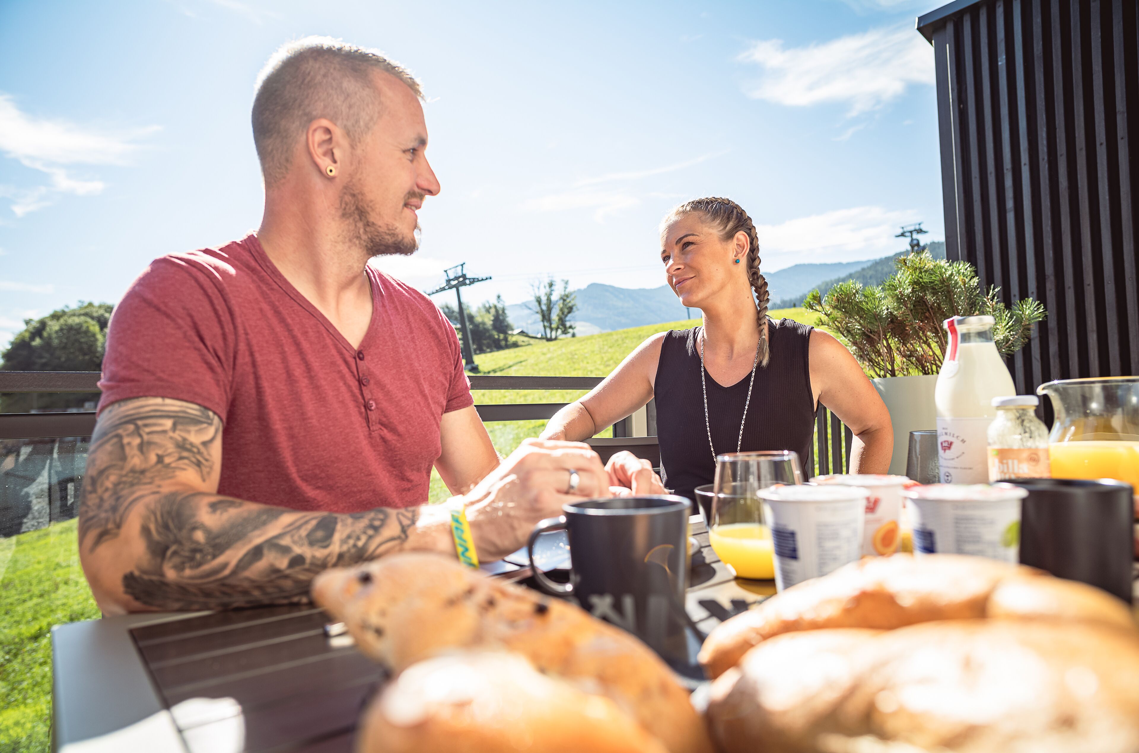 A couple sitting at an outdoor table with breakfast items, overlooking a scenic landscape.