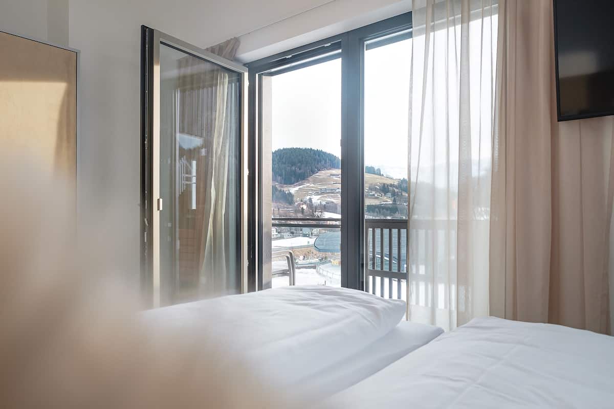 A hotel room with a bed, a sliding glass door leading to a balcony, and a view of a mountainous landscape.