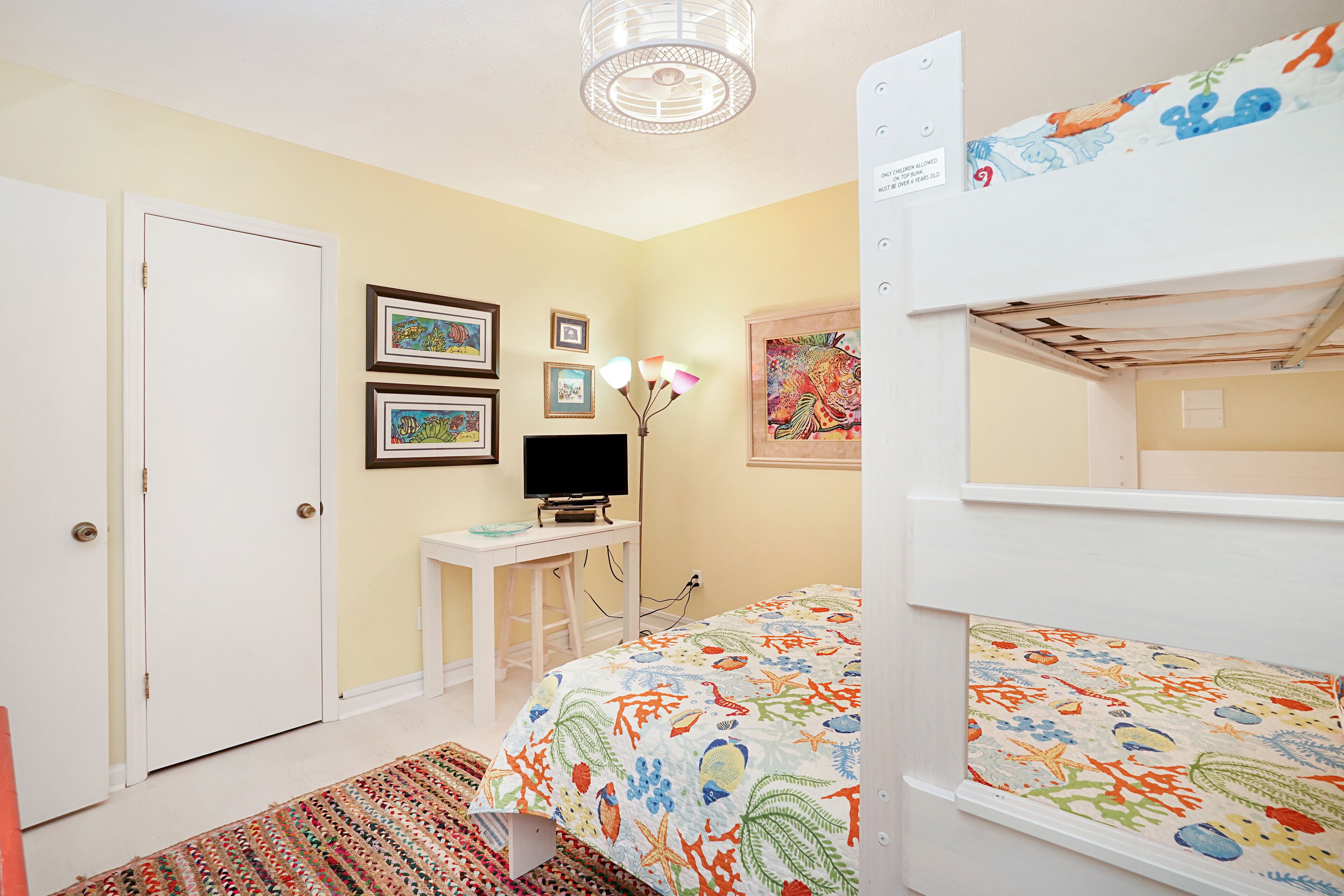 A bedroom with a bunk bed, a desk with a computer, framed pictures on the wall, and a colorful bedspread.